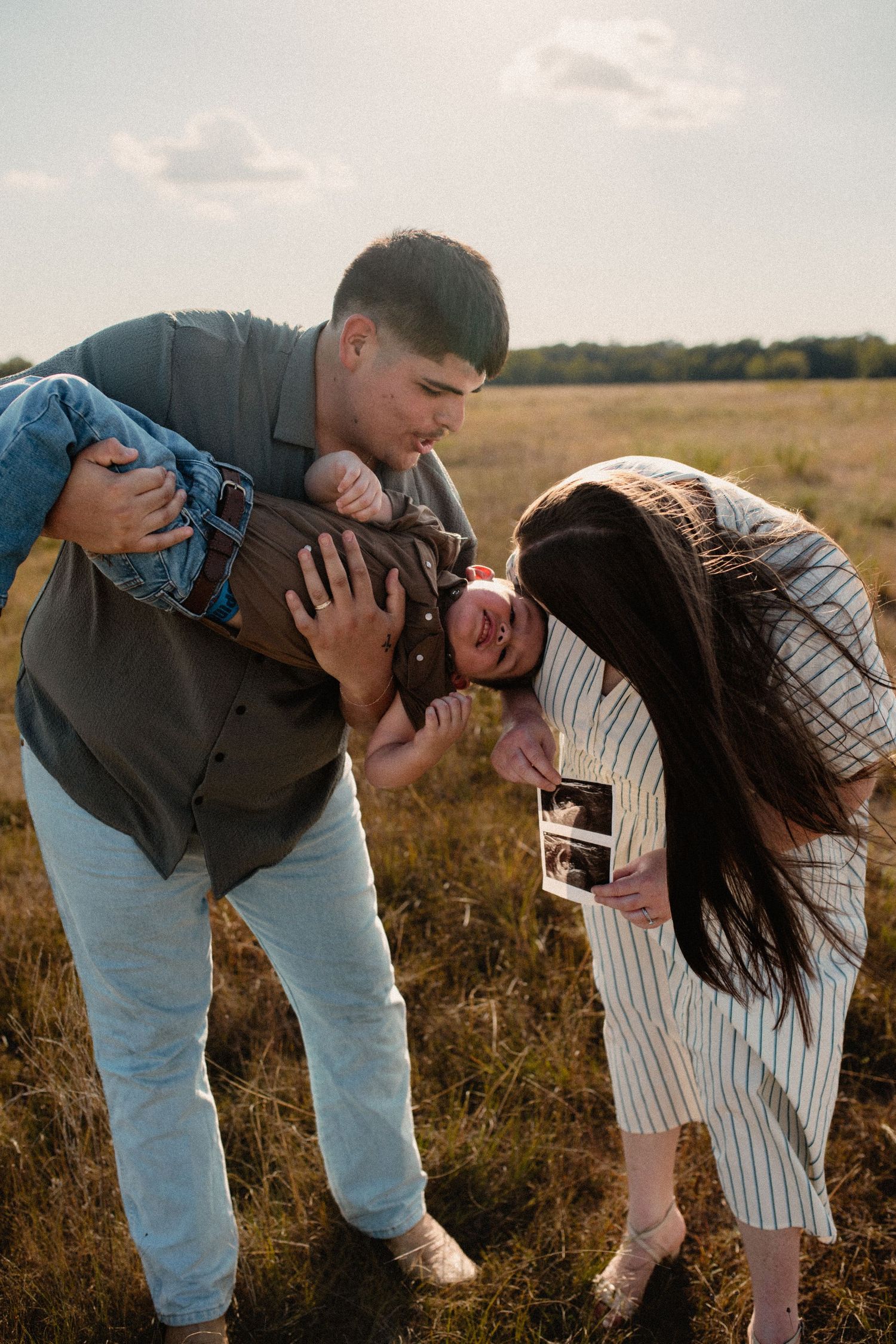 Parents lovingly kiss young boy held by father while mother holds ultrasound in open field.