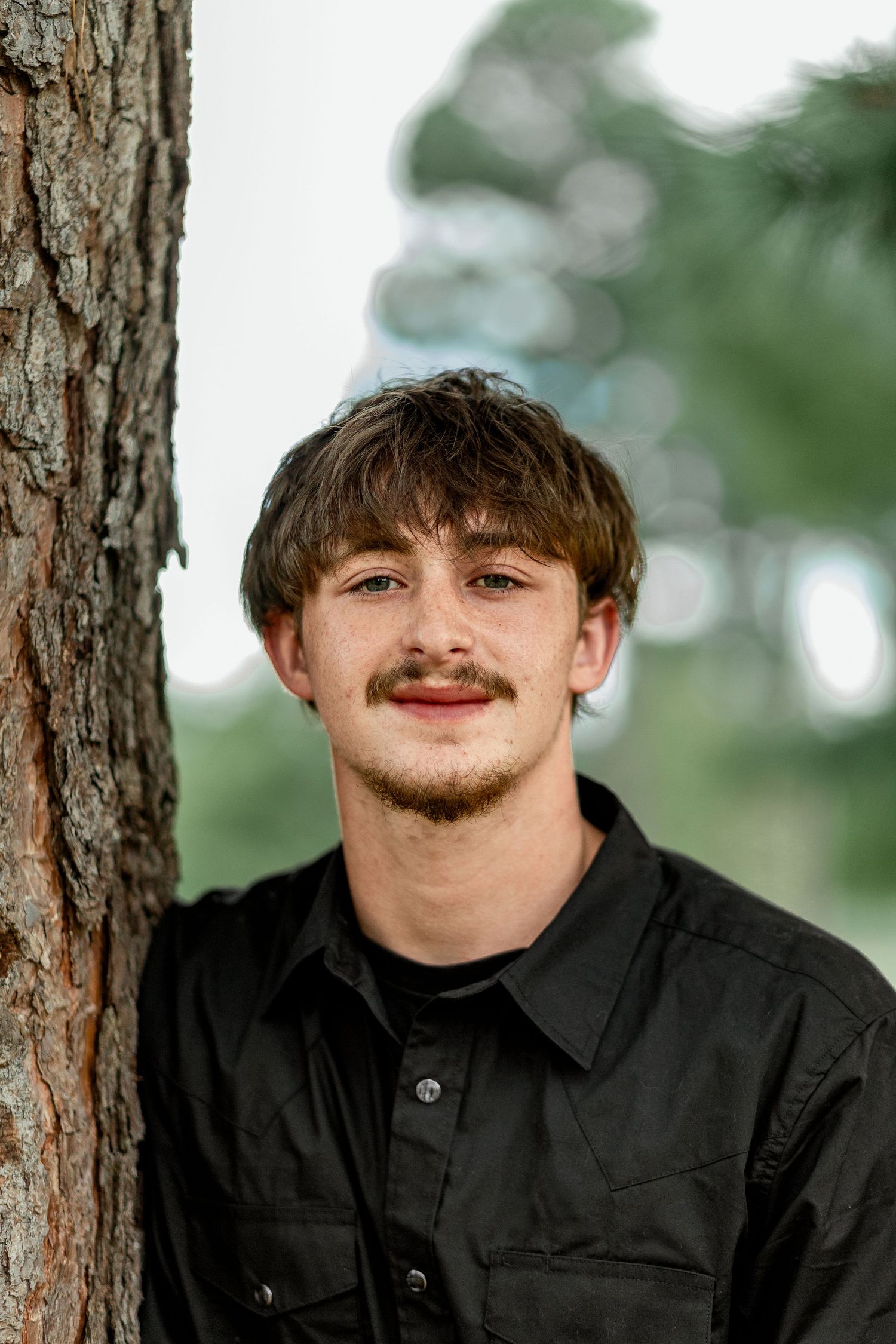 Smiling young man with mustache in black shirt leaning against tree trunk.