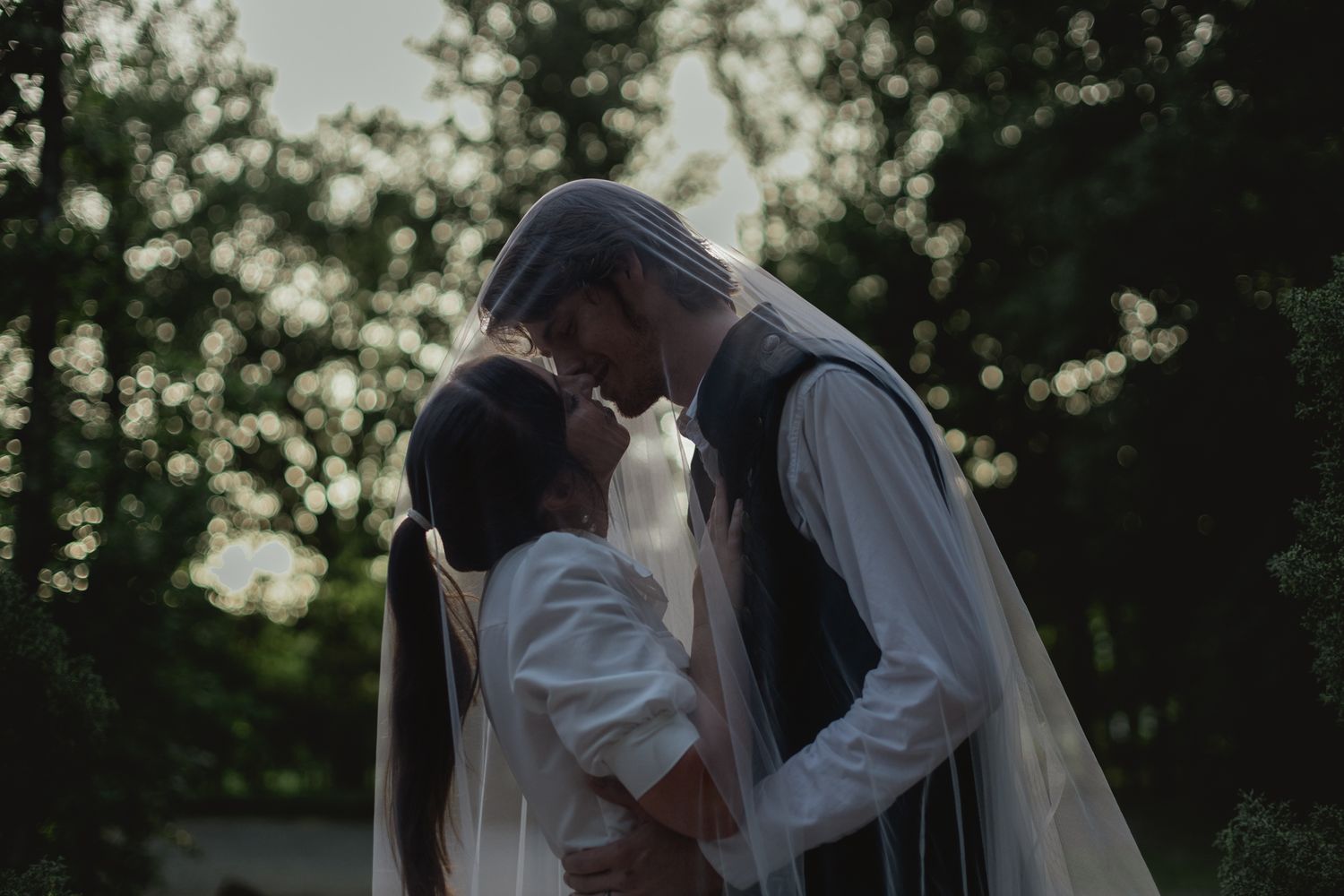 A bride and groom share an intimate moment under a wedding veil surrounded by lush green trees.