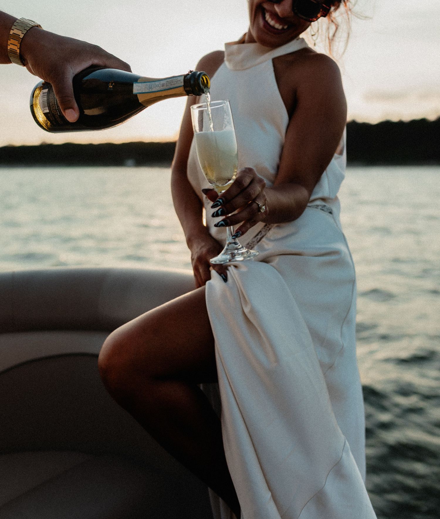 Man pouring champagne into glass held by woman in white dress on boat at sunset.