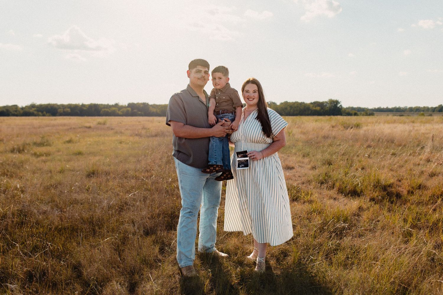 Family of three poses in open field at golden hour, mother holding ultrasound photos.