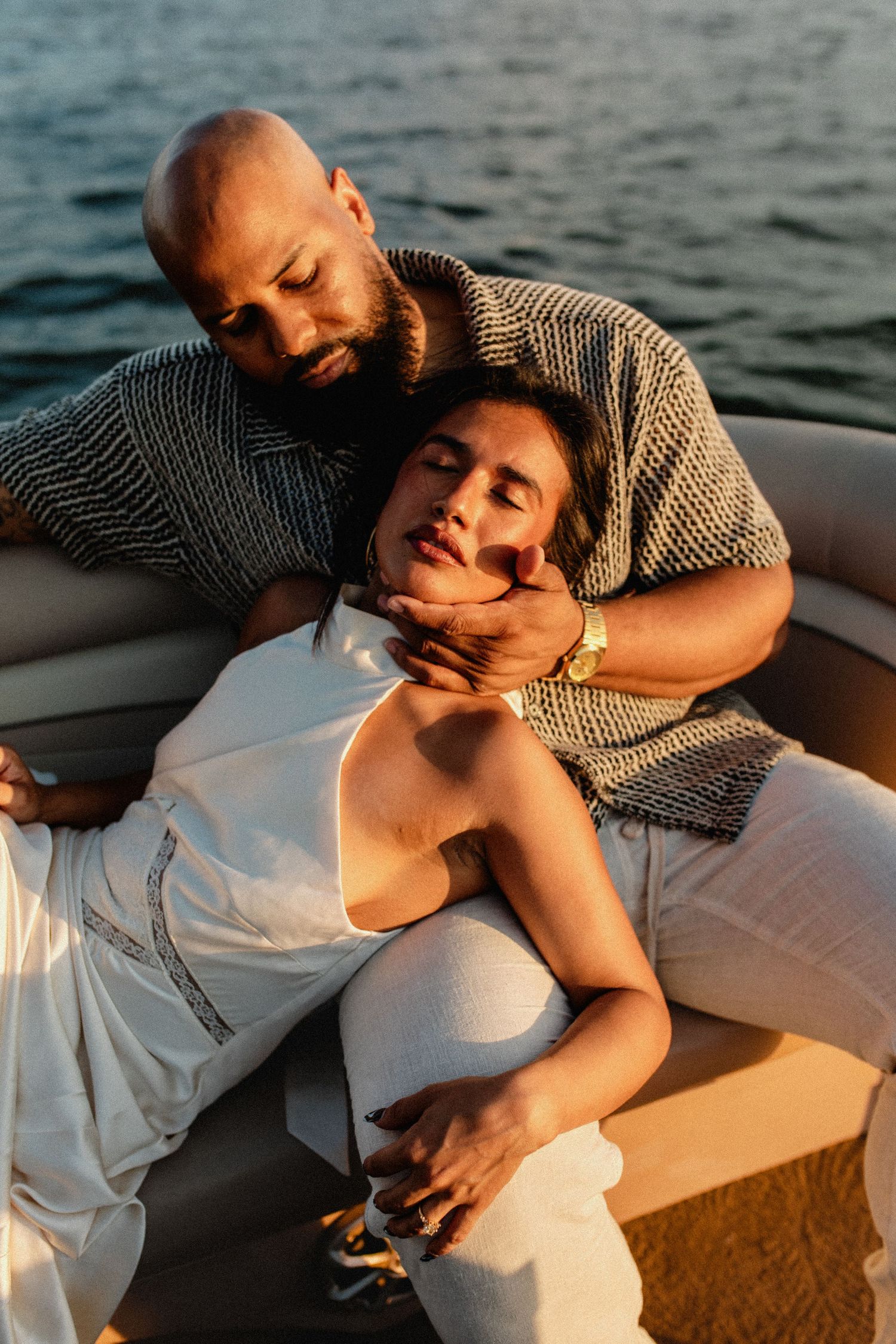 Couple relaxing on a boat at sunset, man embracing woman in white dress near water.