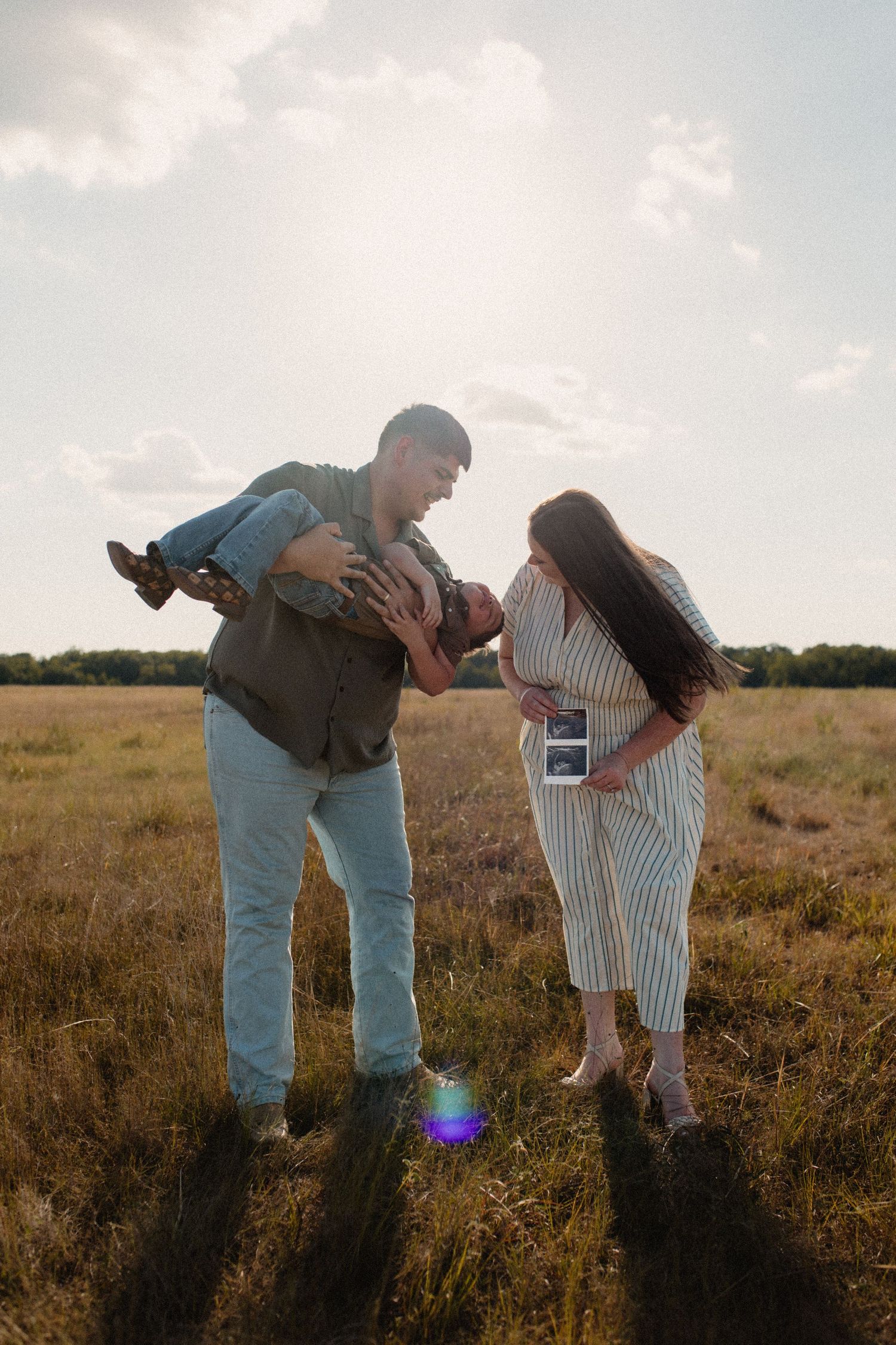 Father playfully holds son as pregnant mother watches, holding ultrasound in sunlit grassy field.