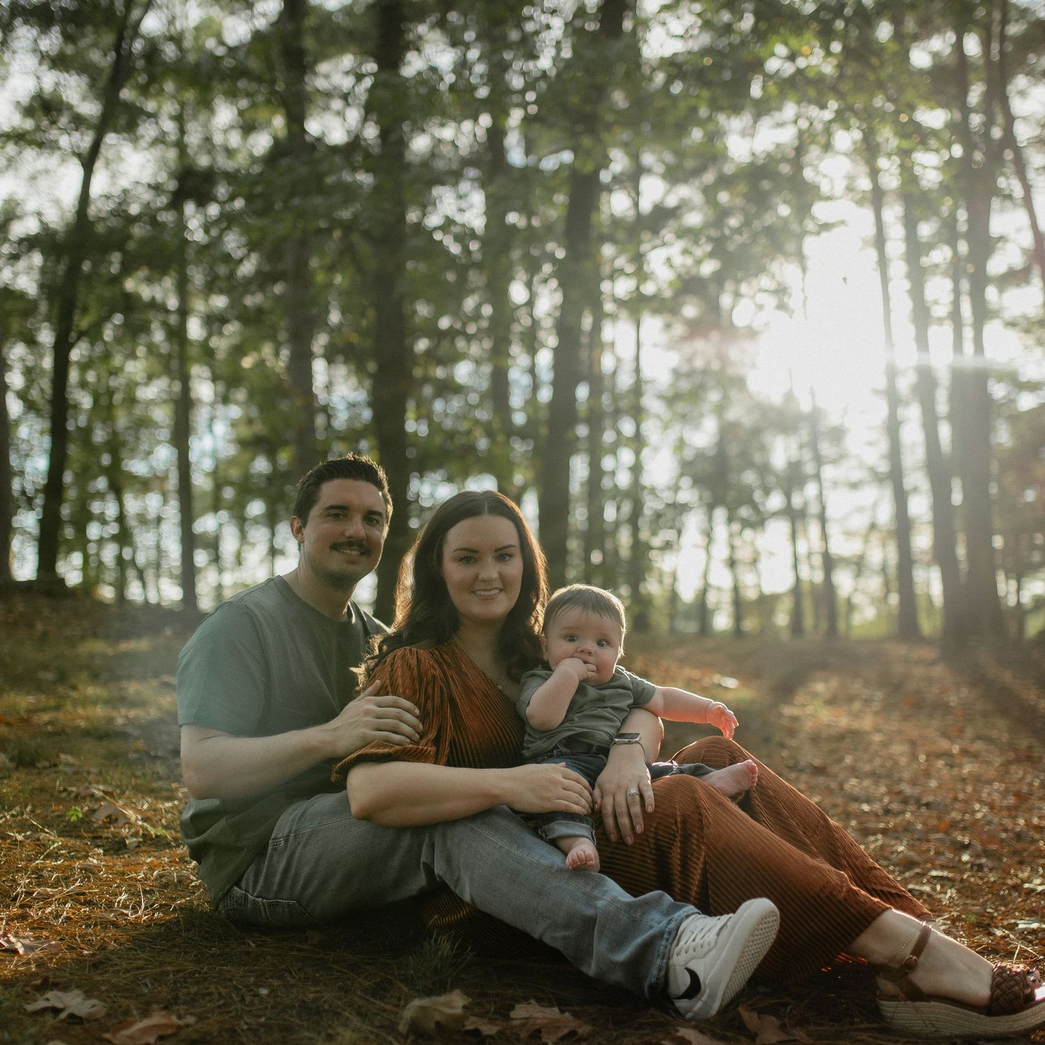 Young family of three sitting together in a sunlit forest, parents smiling with baby on fall leaves.