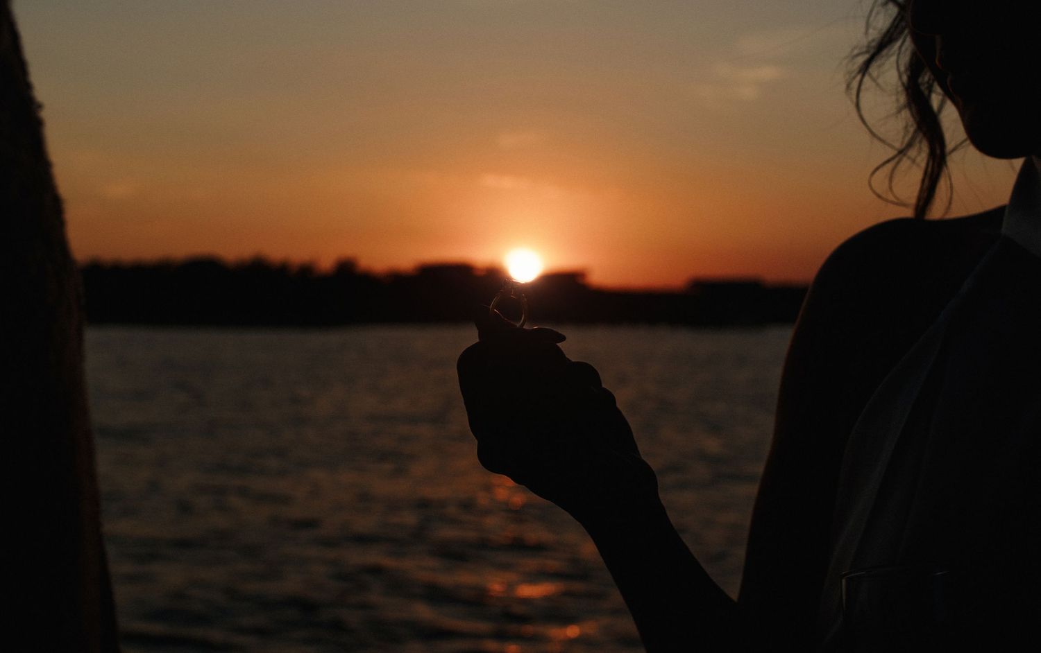 Silhouette of woman holding champagne glass against vibrant orange sunset over calm water.