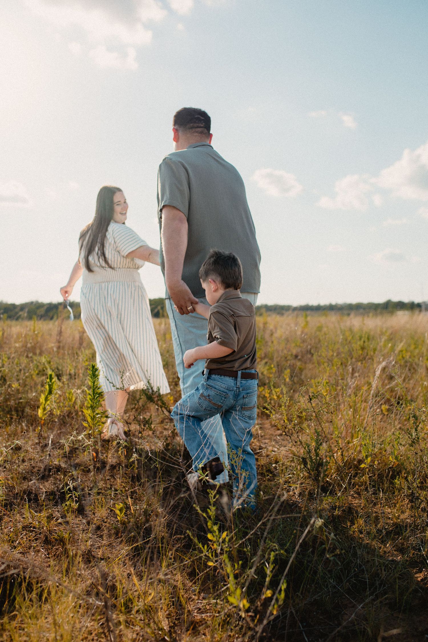 Family of three walking through golden field at sunset, parents holding hands with young boy between them.
