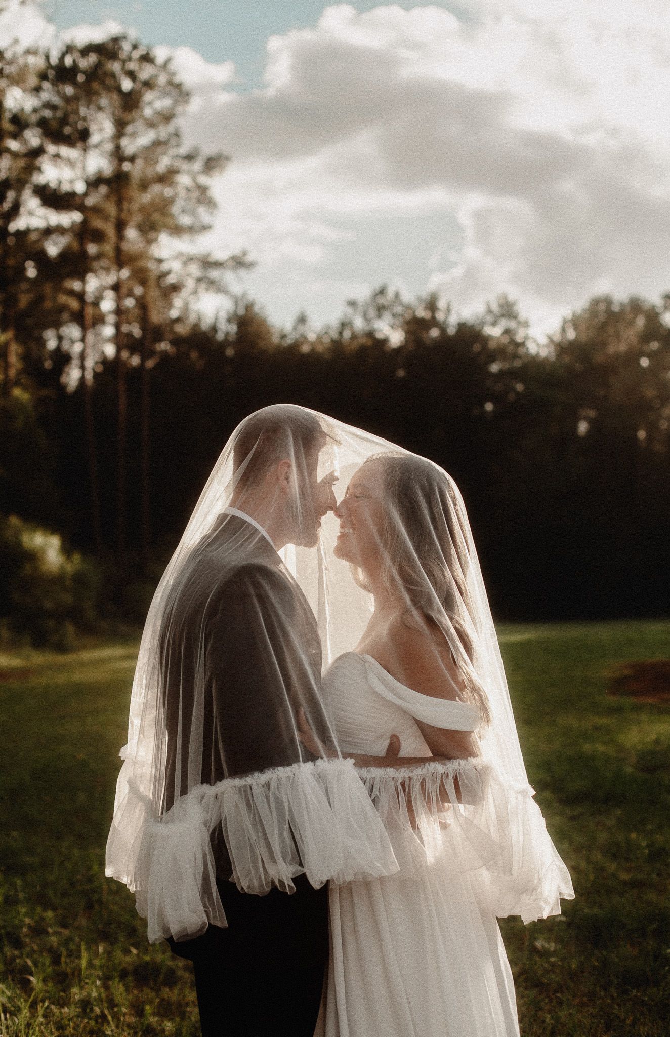 Bride and groom share intimate moment under flowing wedding veil in lush green outdoor setting.