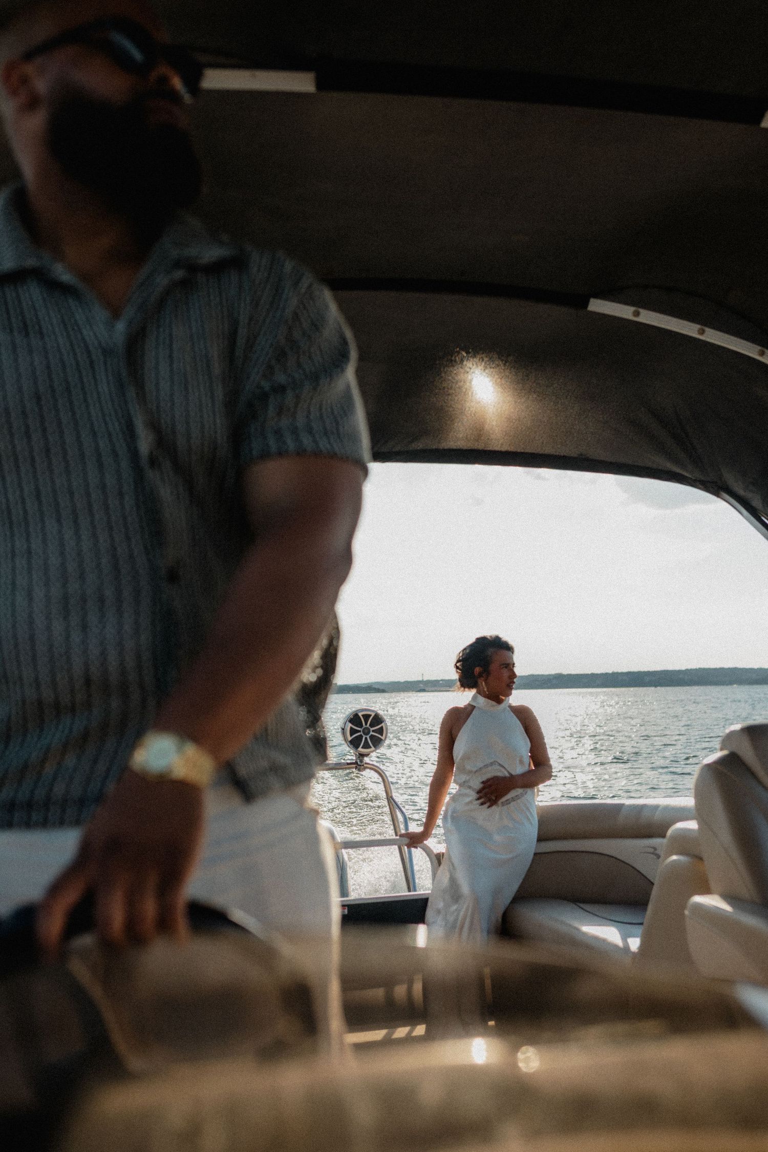 Couple on a luxury boat, man in striped shirt with gold watch, woman in white halter dress standing near water.