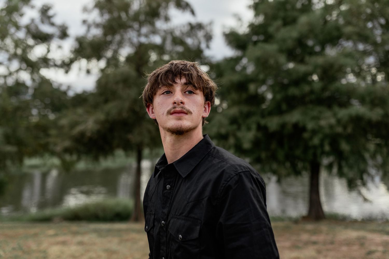 Young man with curly hair and mustache wearing black shirt, looking up outdoors among green trees.