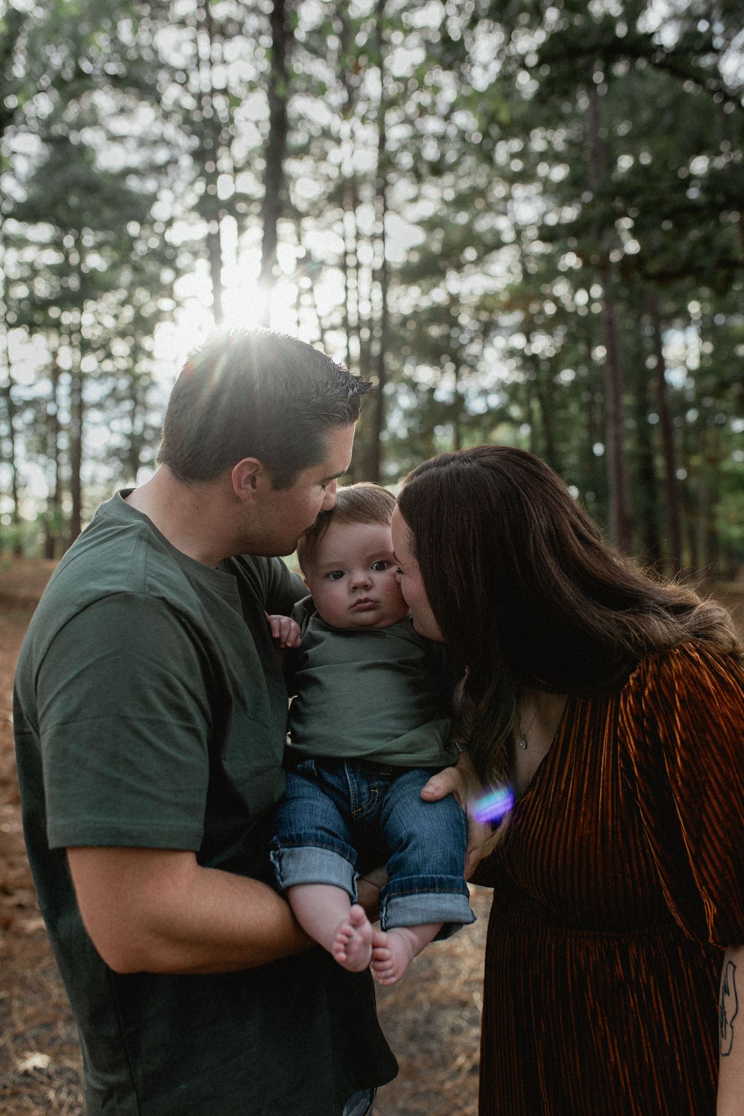 Parents lovingly kissing their infant on the cheeks in a pine forest at sunset.