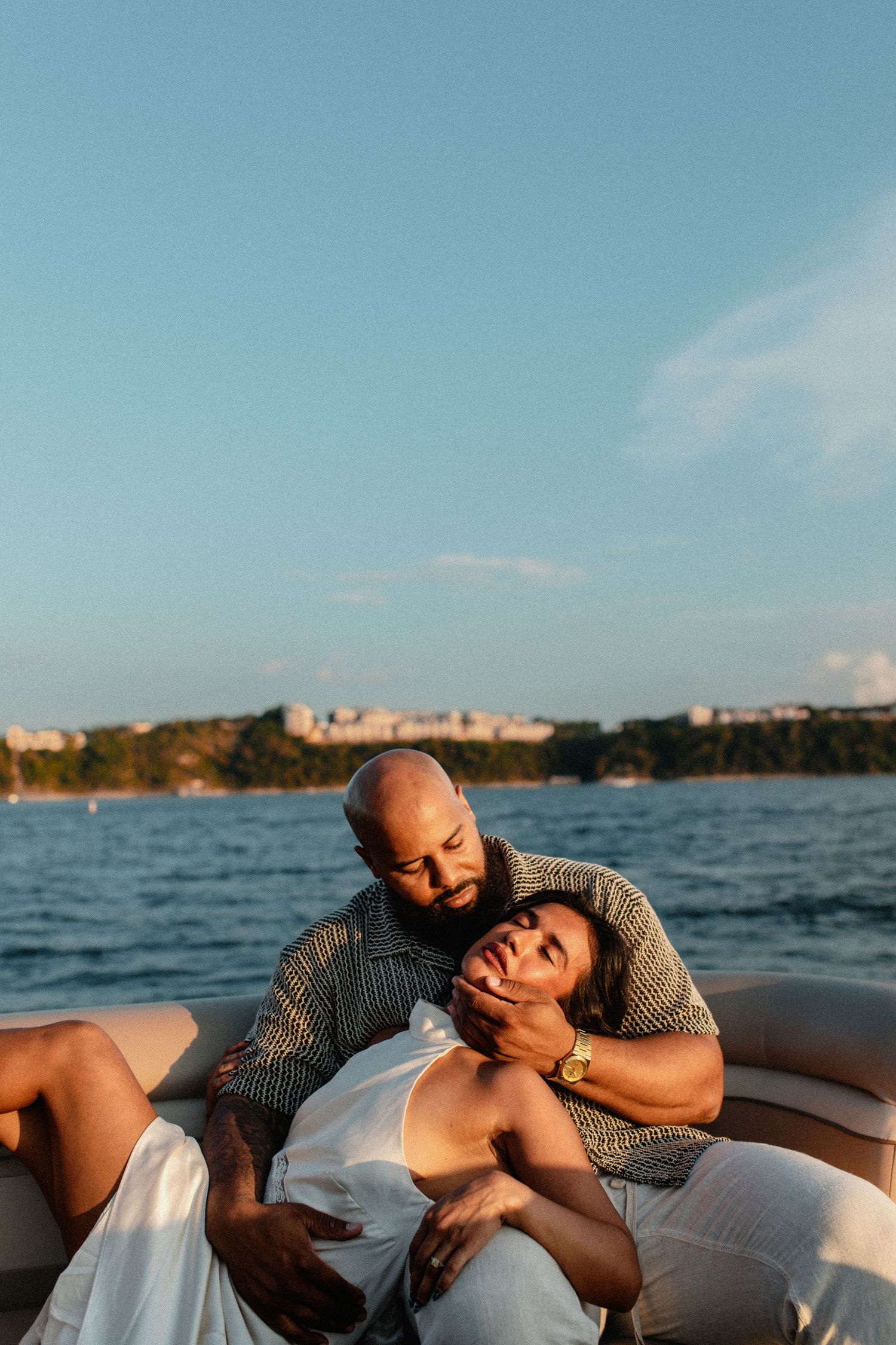 Couple relaxing on boat, woman reclining on mans lap with cityscape and blue water behind them.