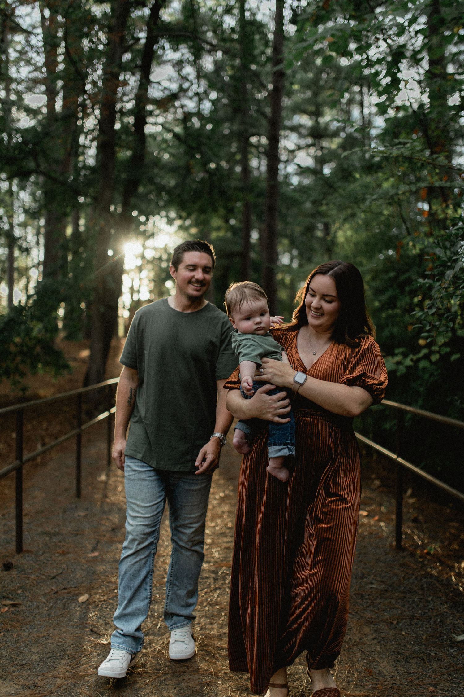 A happy family walks along a sunlit forest path, with the mother carrying their baby boy.