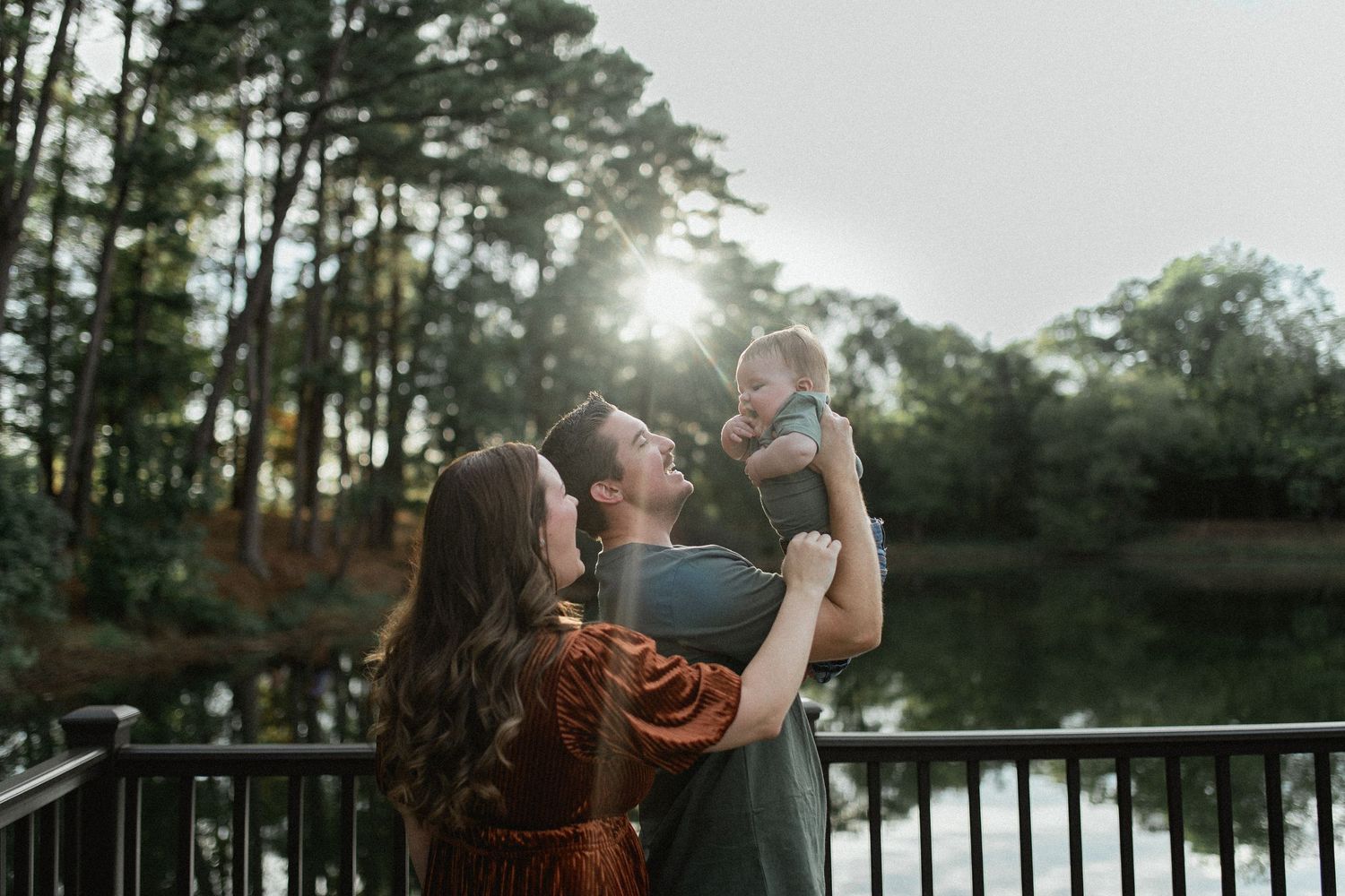 A couple lifts their baby in golden backlight by a lake, surrounded by lush green trees.