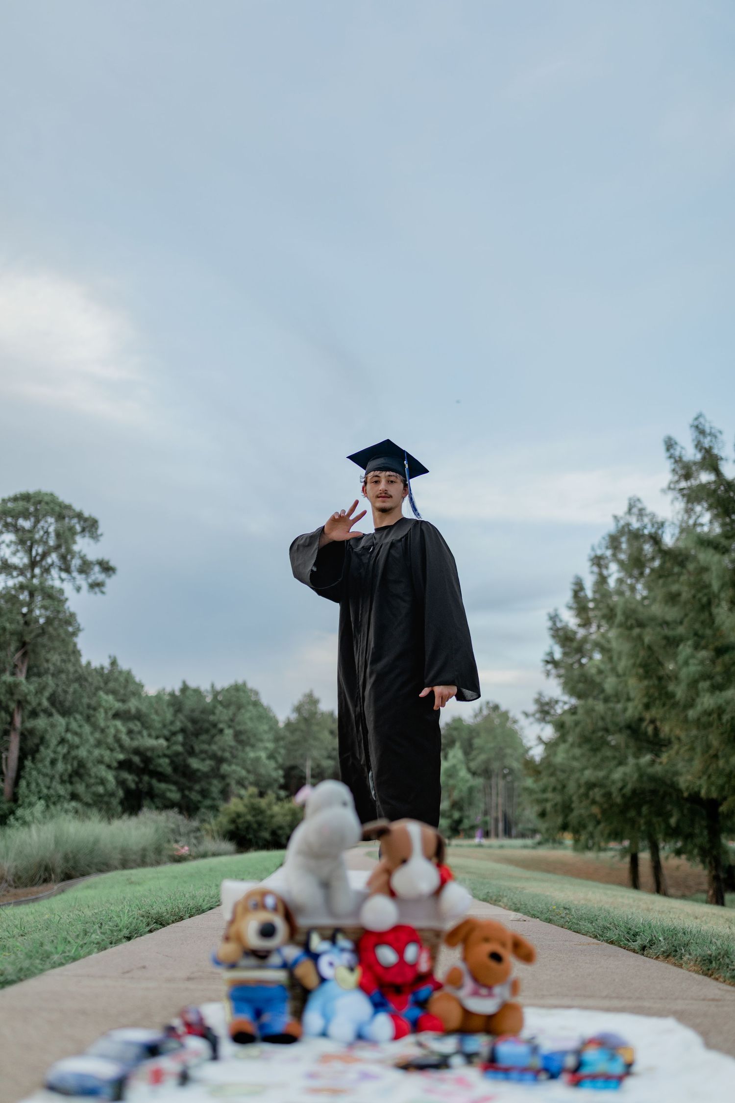 Graduate in cap and gown stands on pathway surrounded by childhood toys and stuffed animals outdoors.