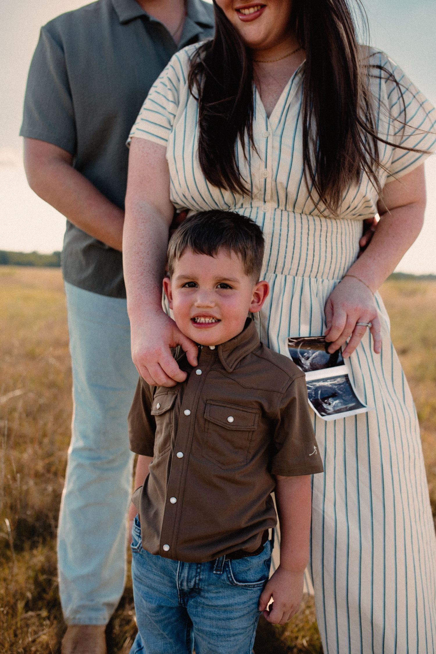 Smiling boy stands between parents holding ultrasound photo during family pregnancy announcement session.