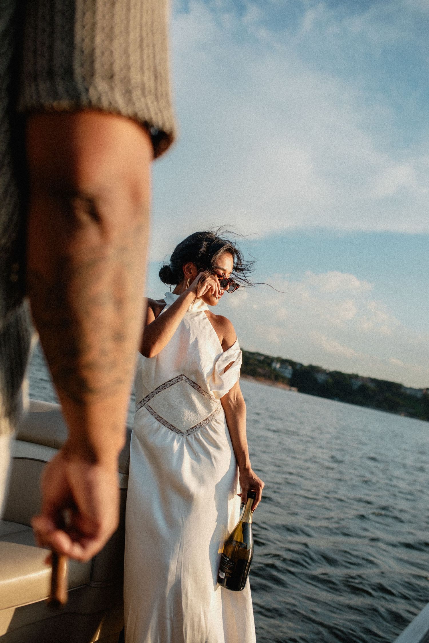 Woman in white dress holding champagne bottle on a yacht at golden hour with wind in her hair.