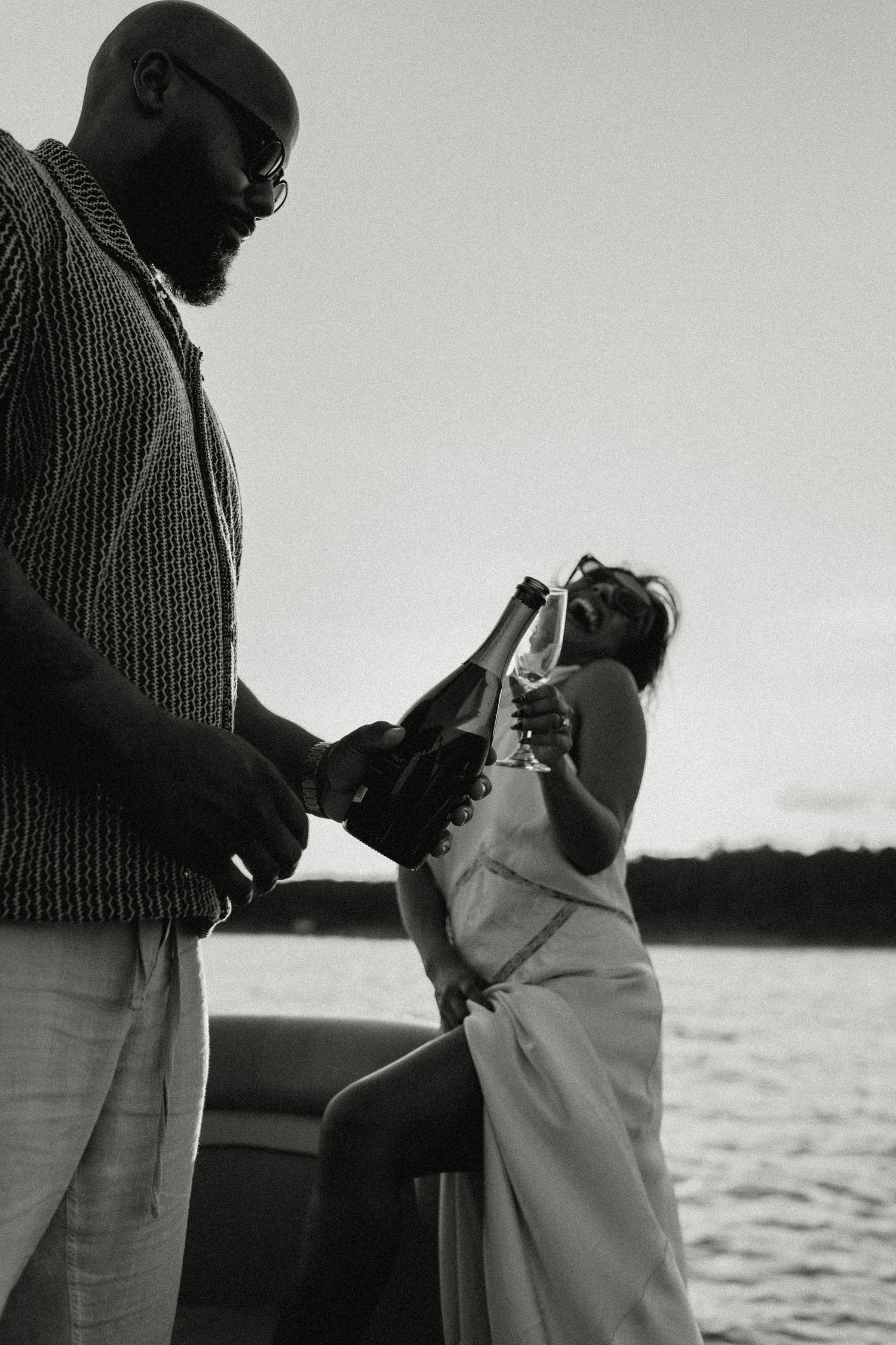 Black and white image of man pouring champagne for woman in white dress on boat.