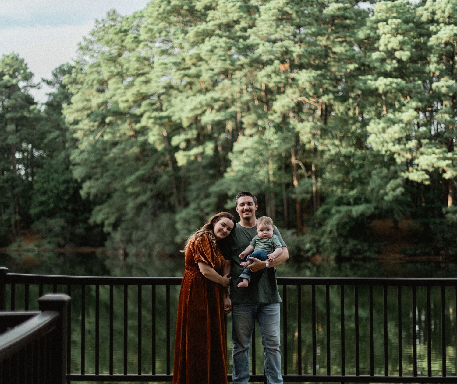 Happy young family with baby posing on a bridge surrounded by lush green trees near a pond.