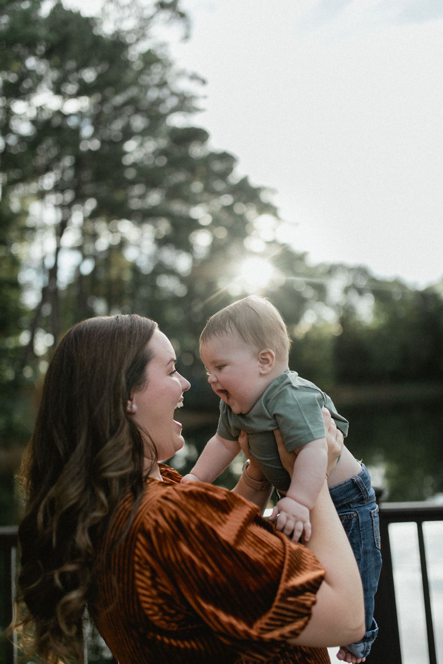 A laughing mother lifts her giggling baby in golden sunlight near a lakeside railing outdoors.