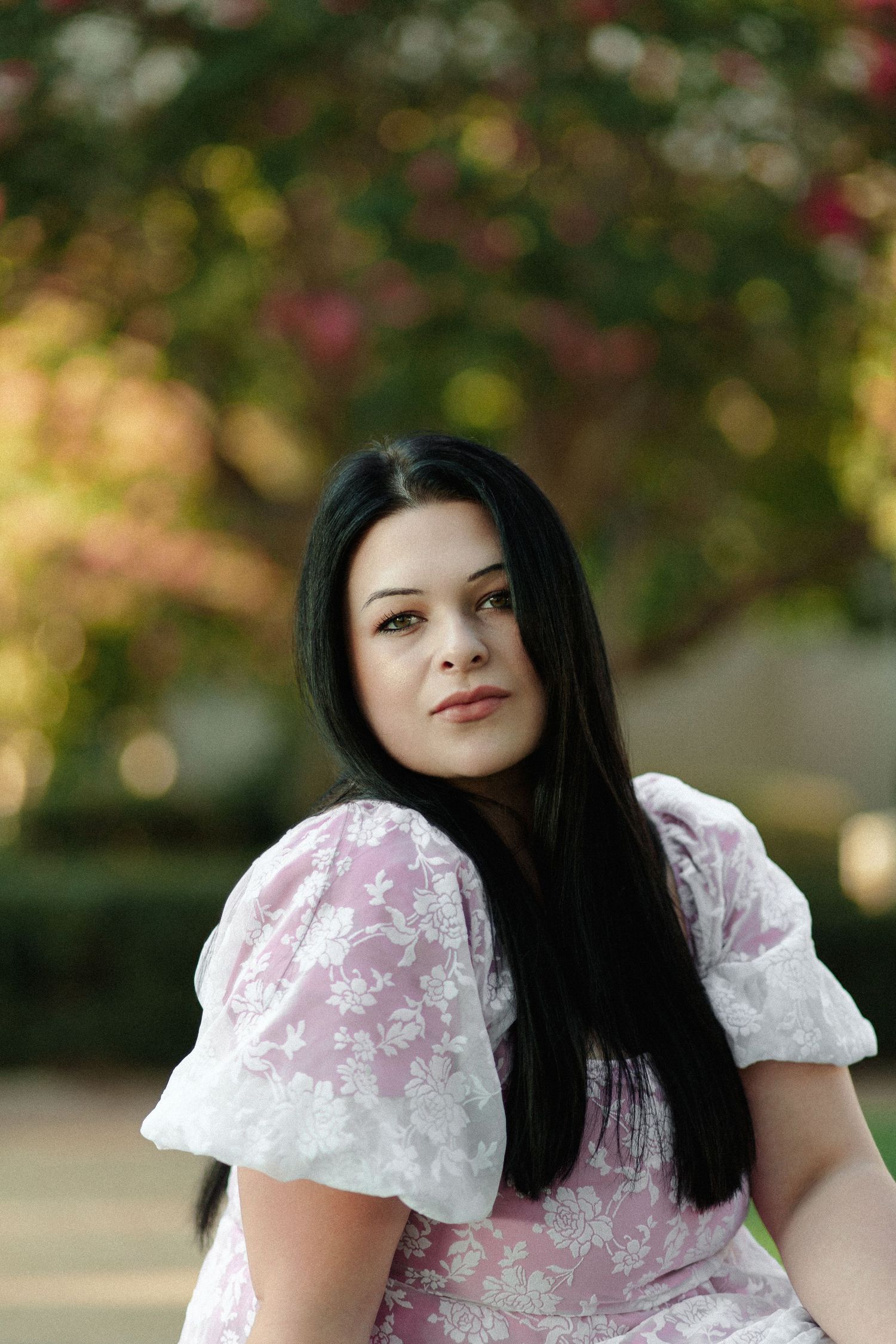 Young woman with long black hair wearing a pink floral dress poses outdoors near blooming trees.