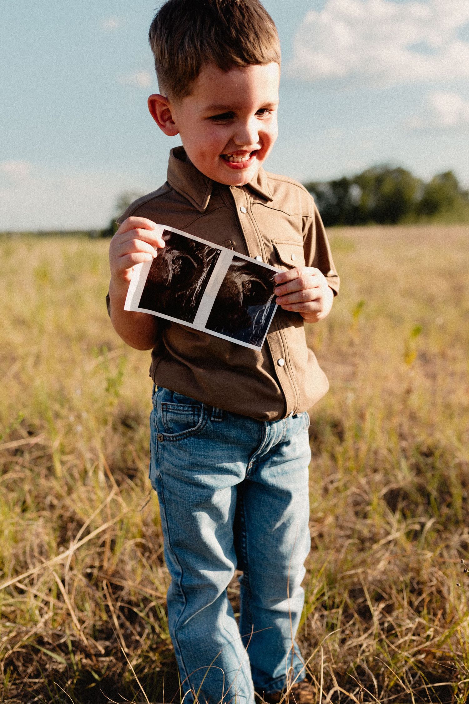 A smiling boy in a brown shirt holds an ultrasound photo in a sunny grassy field.