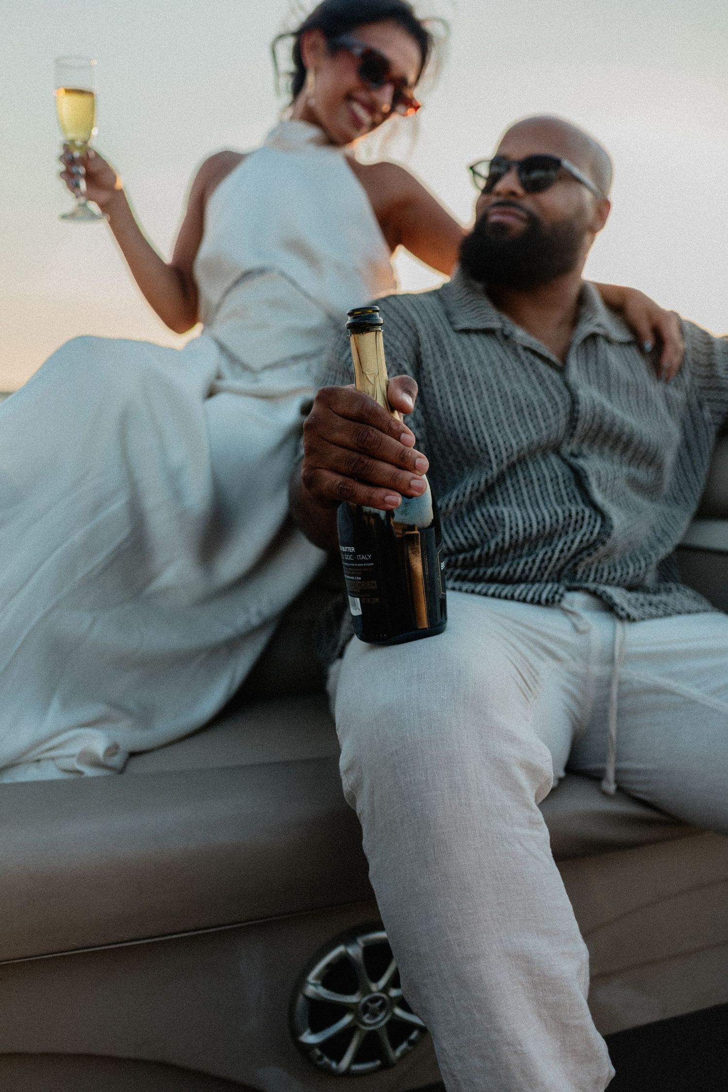 Couple relaxing on boat at dusk, man holding champagne bottle, woman raising glass.
