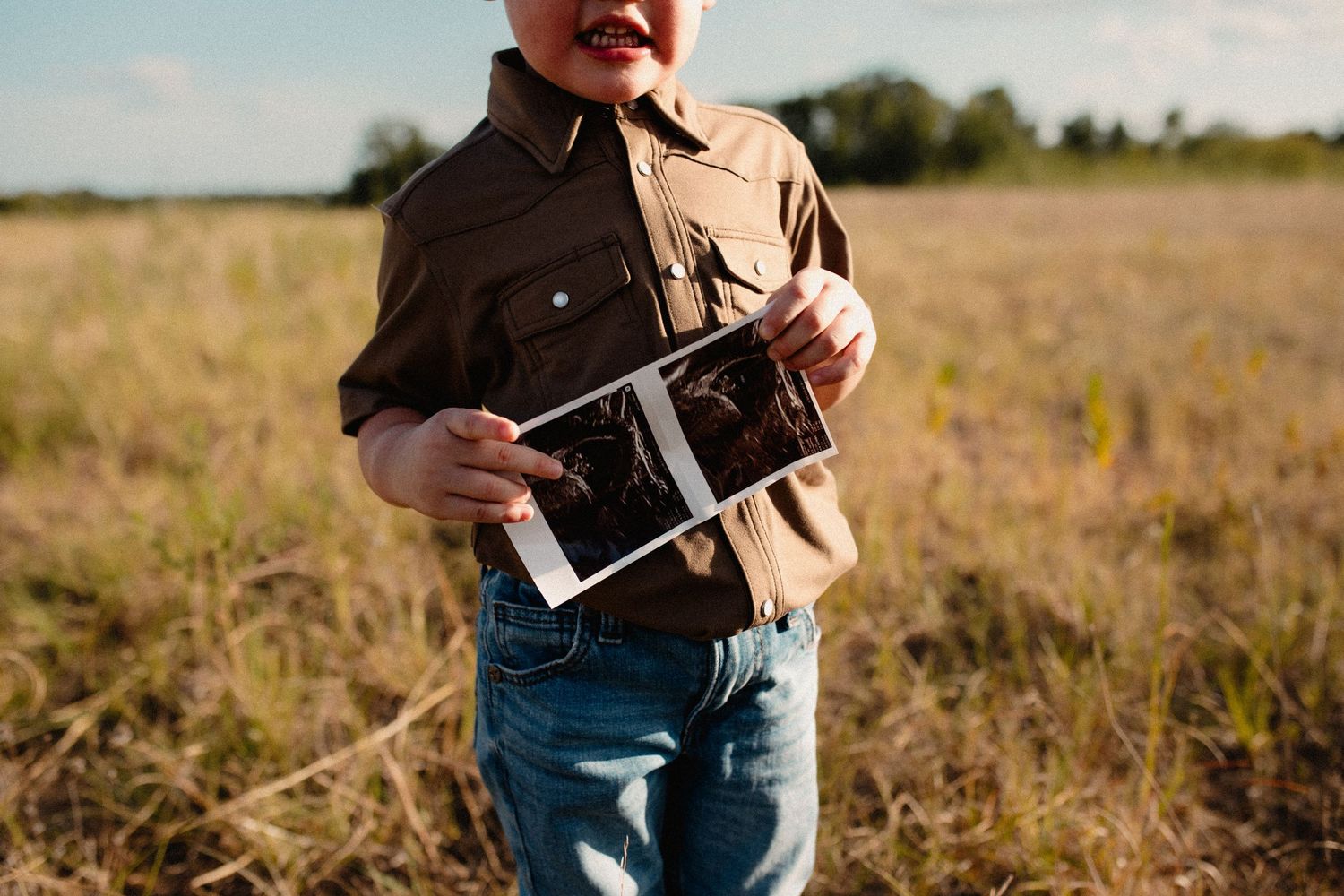 Close-up of a child in a brown shirt proudly displaying an ultrasound photo outdoors.