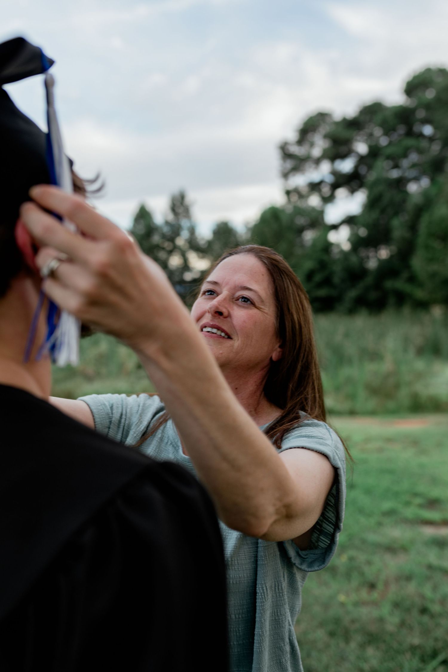 Smiling woman adjusting graduation cap outdoors with trees in background.