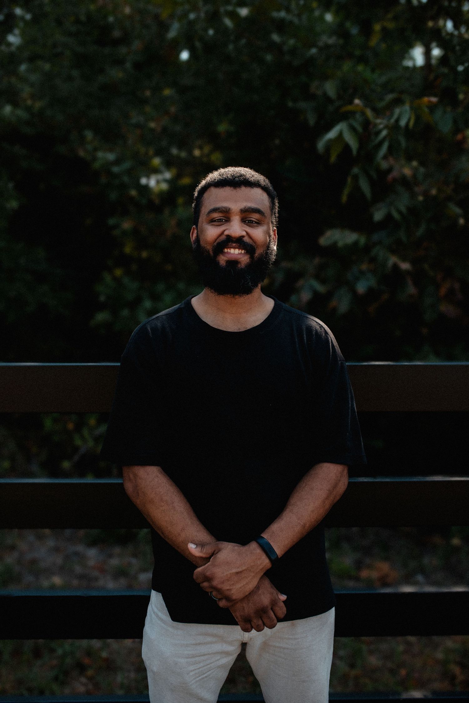 Smiling bearded man in black t-shirt stands confidently before a dark wooded background.