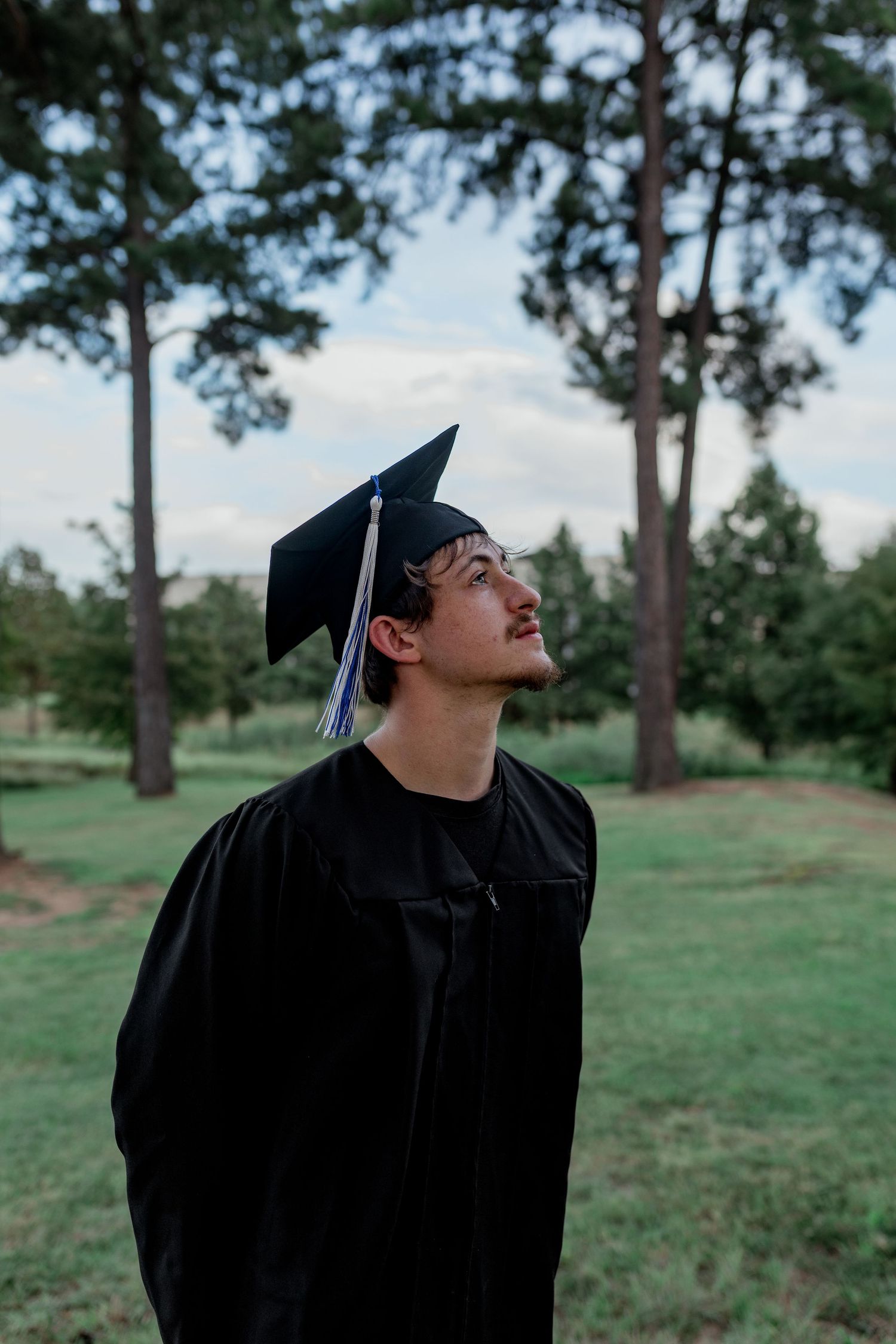 Young male graduate in black cap and gown looks upward outdoors among green trees.