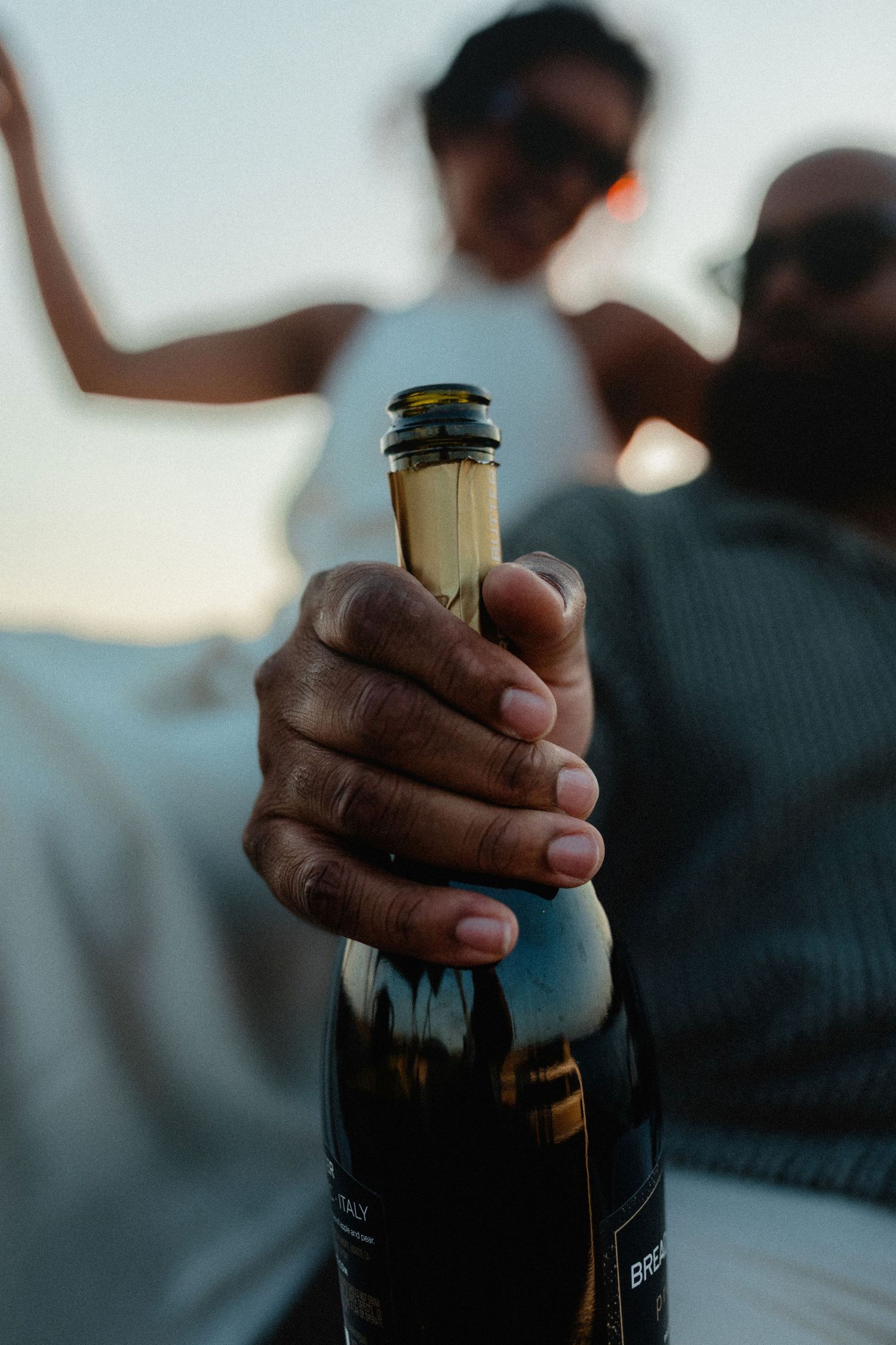 Close-up of hand holding champagne bottle toward camera with couple relaxing on boat in background.