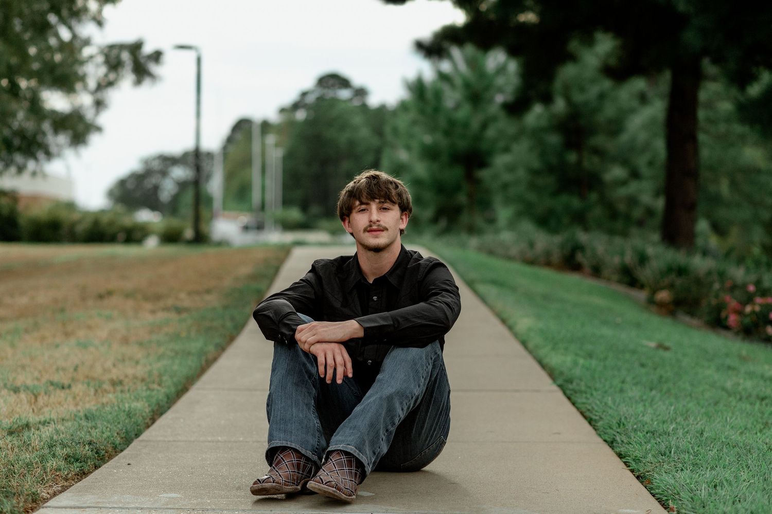 Young man in black shirt sitting cross-legged on sidewalk in green park.