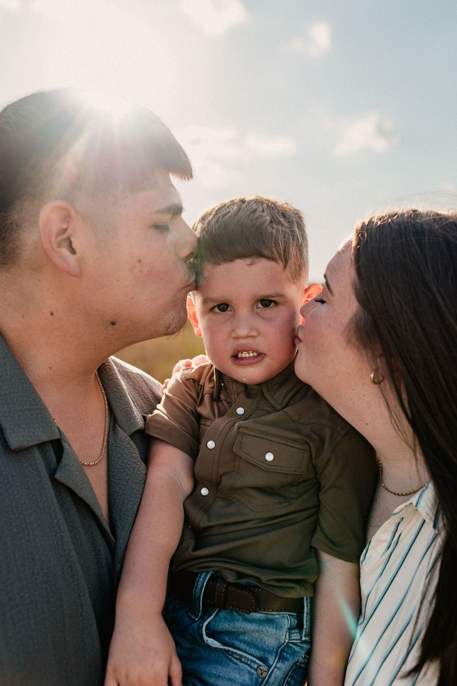 Parents simultaneously kiss their young son on both cheeks in a sunny outdoor field.