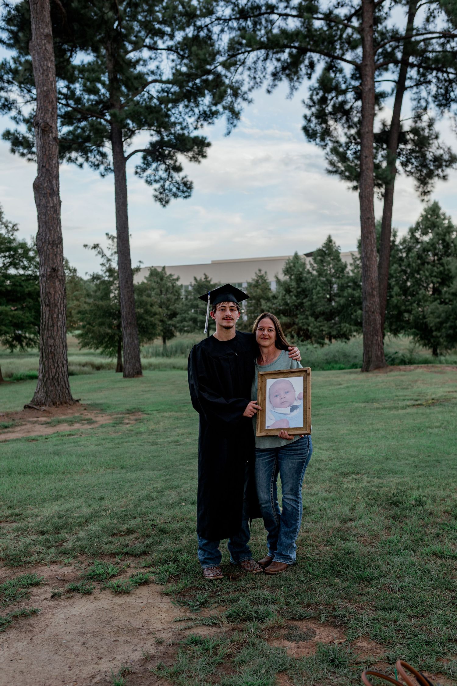 Graduate in cap and gown poses with woman holding framed baby photo outdoors among tall pine trees.