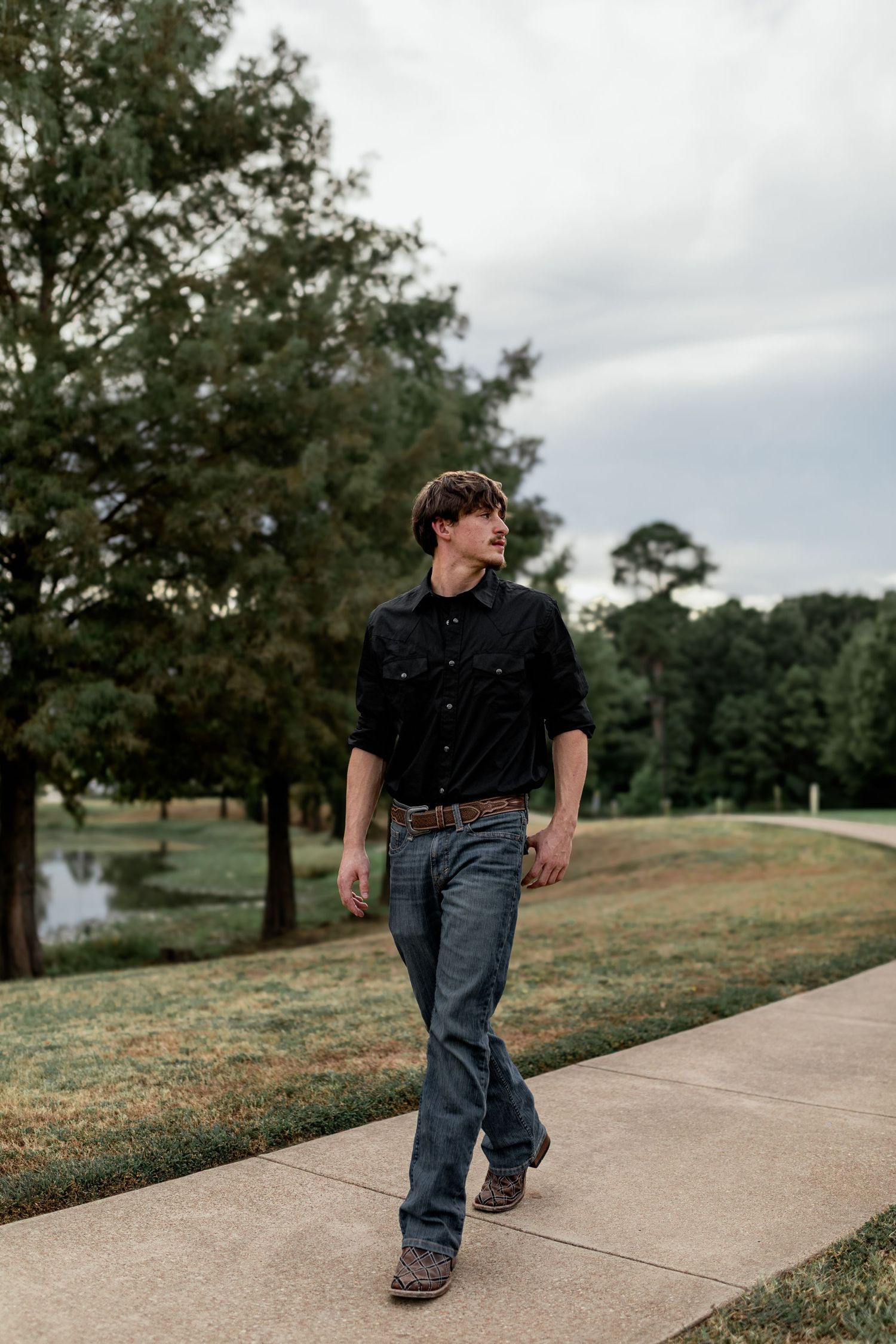 Young man in black button-up shirt and jeans walking confidently along a sidewalk in a green park.