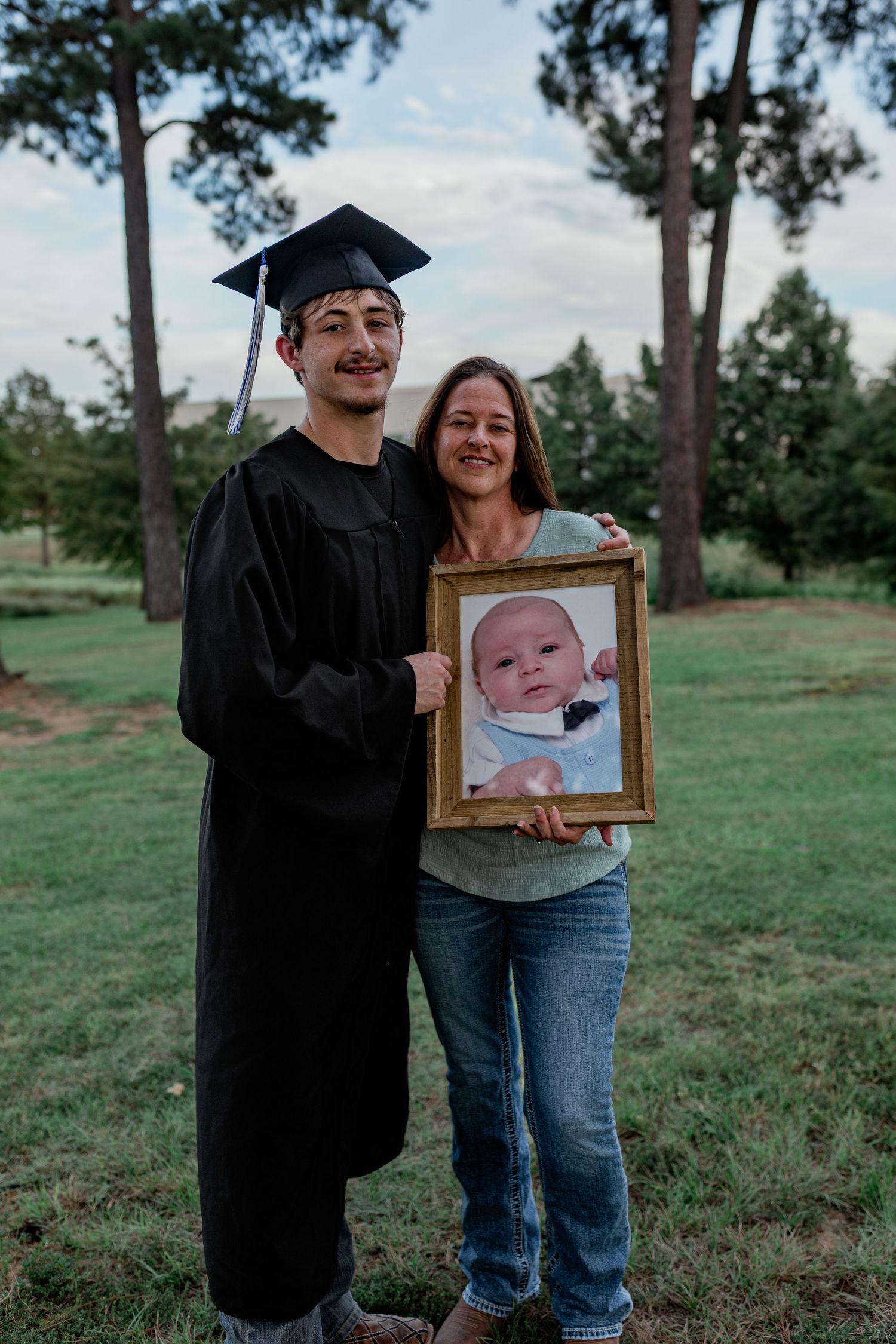 Graduate in cap and gown holding framed baby photo with smiling woman outdoors.
