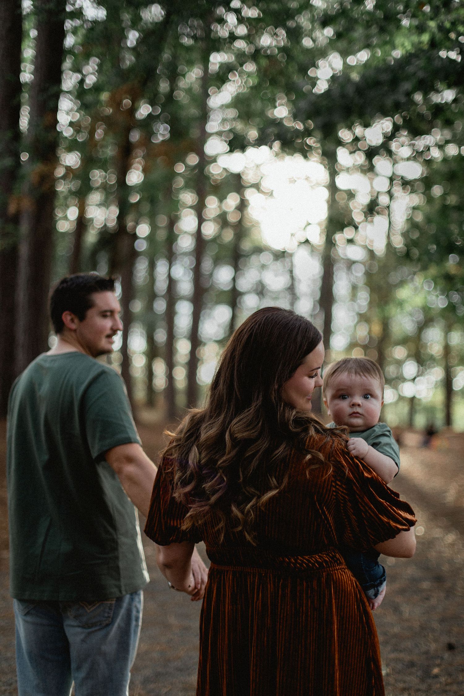 A mother with long wavy hair holds her baby while a man stands nearby in a sunlit pine forest.