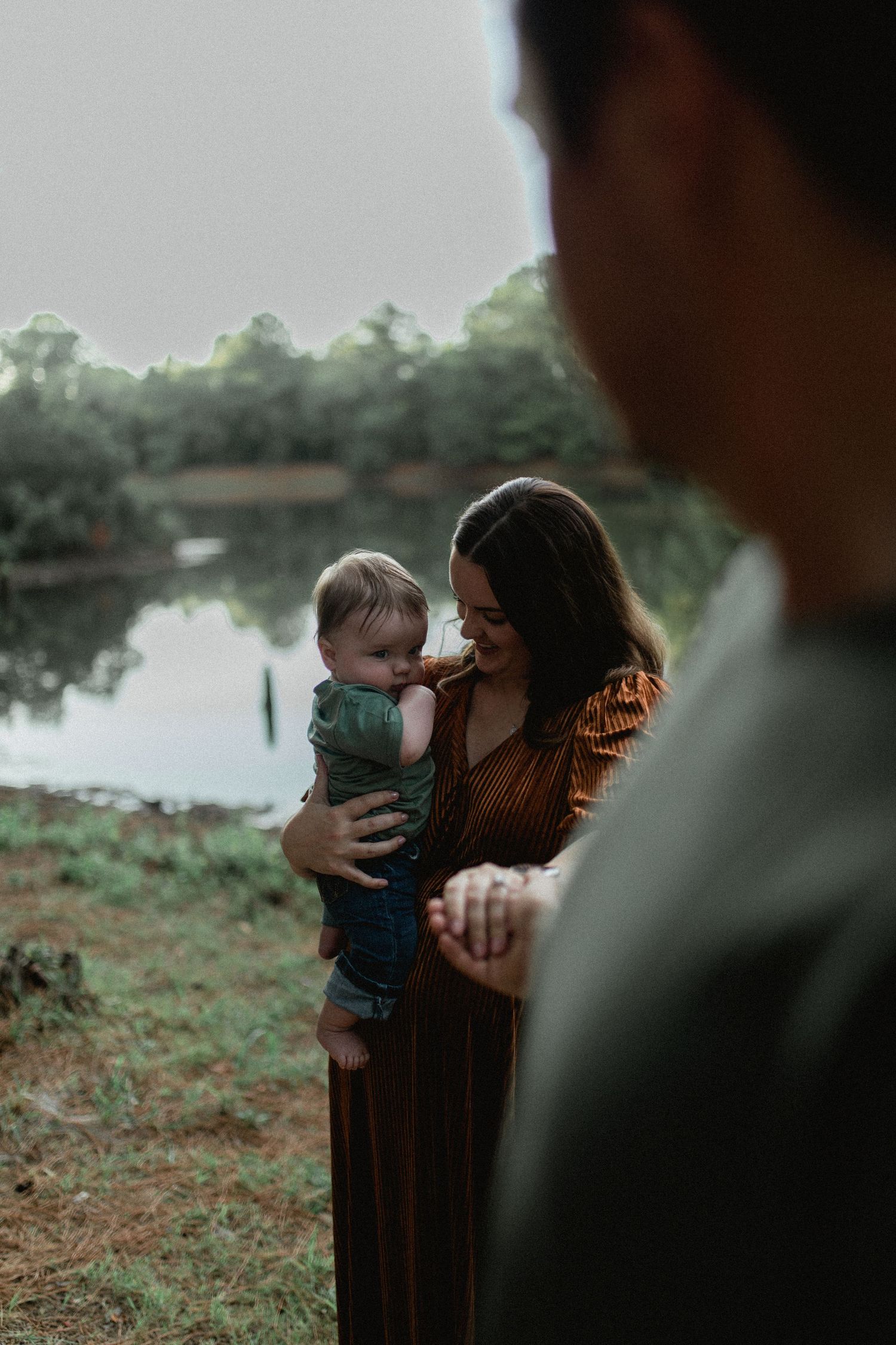 Mother holding baby near a serene lake, smiling tenderly while father stands in foreground.