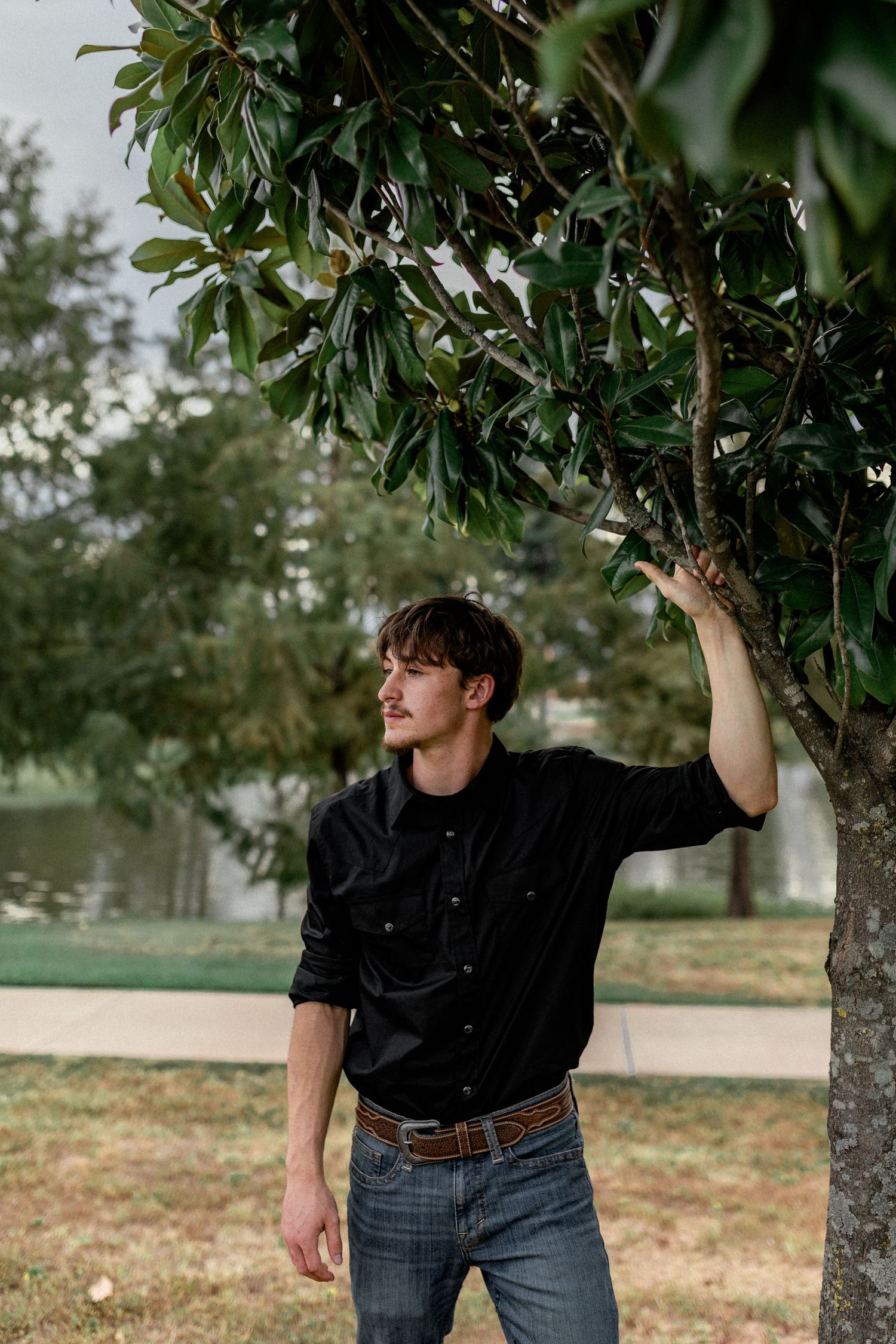 Young man in black shirt standing beside magnolia tree gazing away in outdoor park setting.