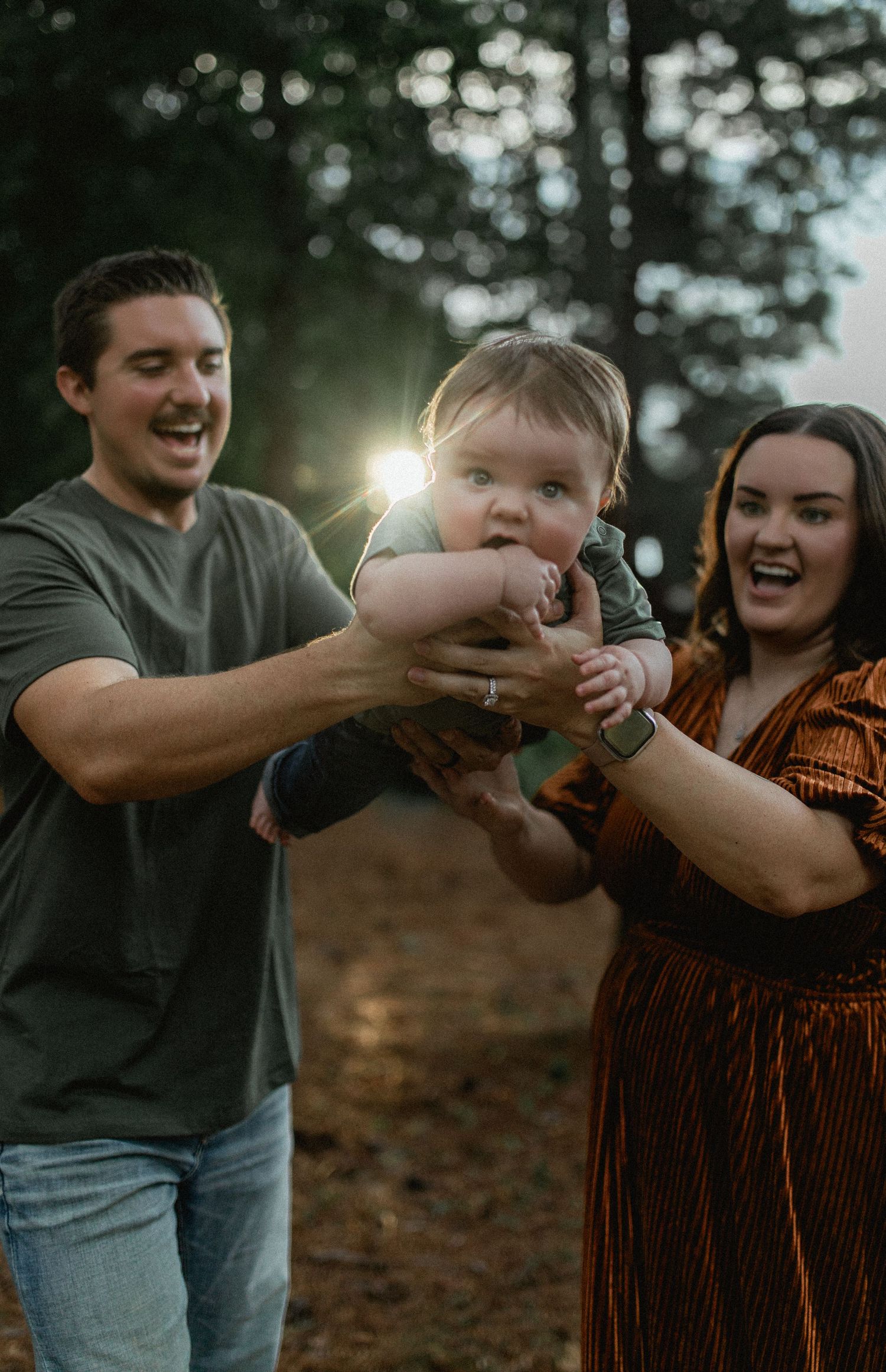 Laughing couple holding up their cute baby outdoors in golden hour forest light.