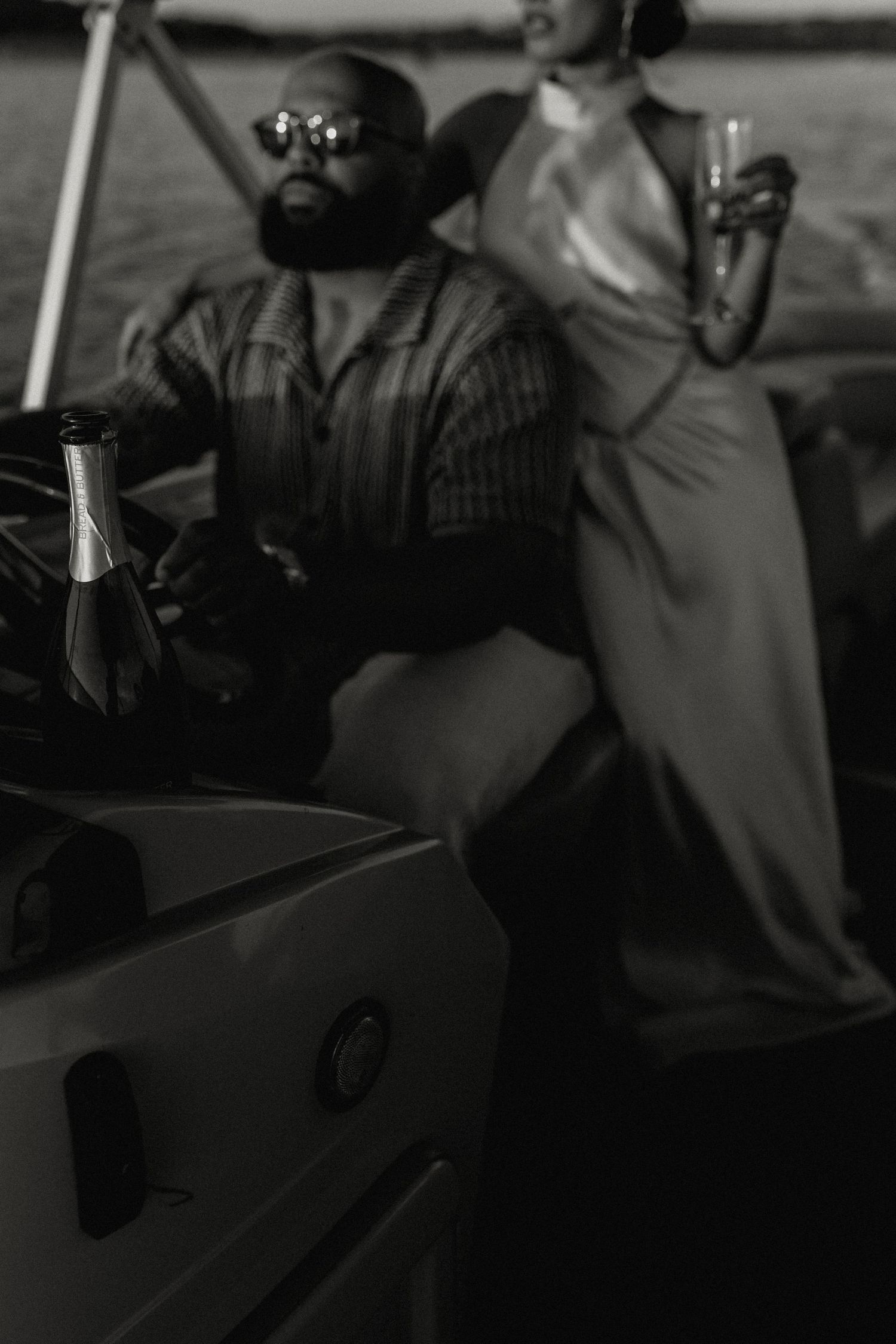 Black and white photo of couple on boat with champagne bottle in foreground at dusk.