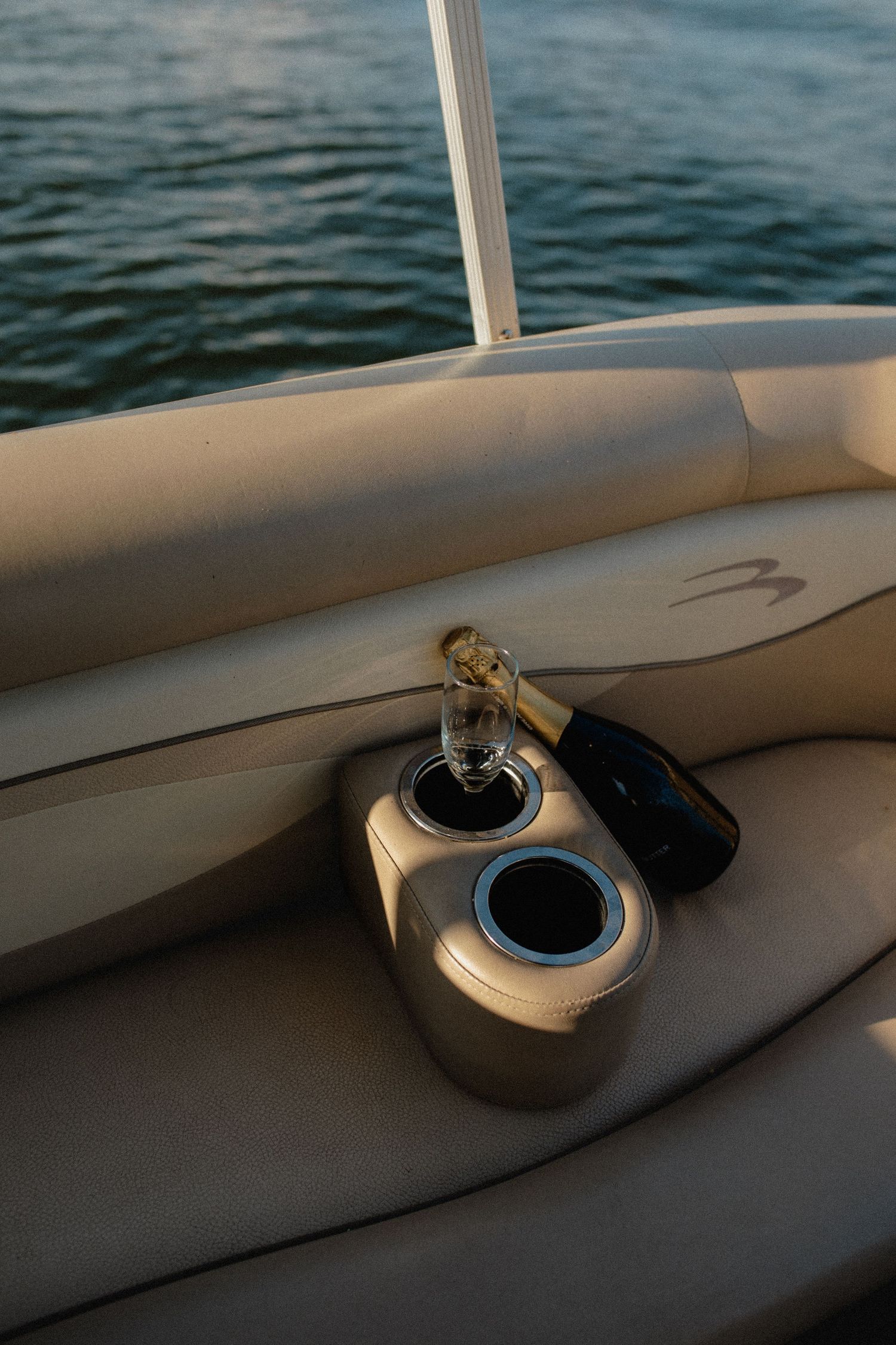Champagne bottle and glass in boat cup holder on beige leather seat with serene water in background.