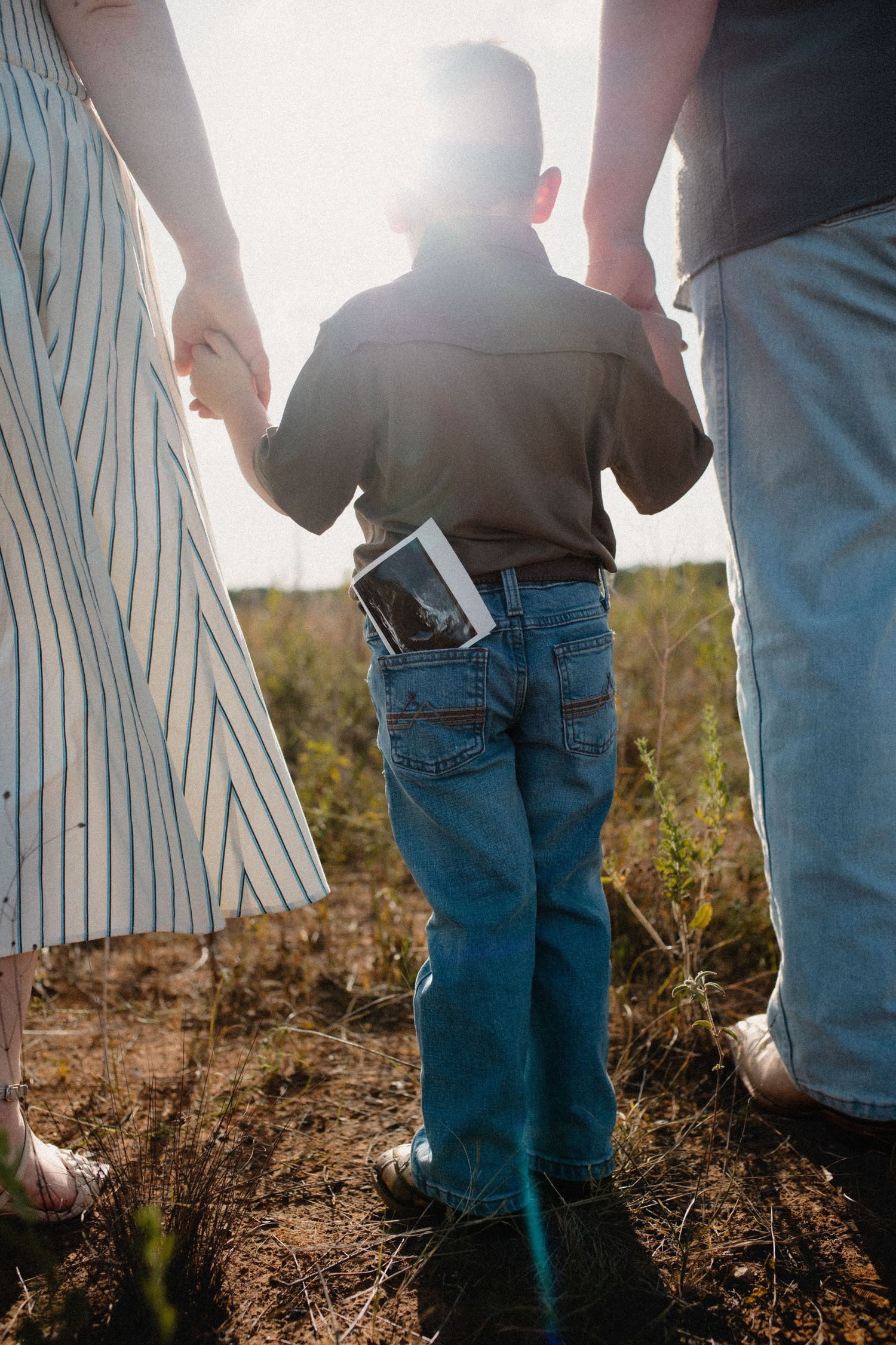 Child holding ultrasound photo while walking hand-in-hand with parents in sunny outdoor field.