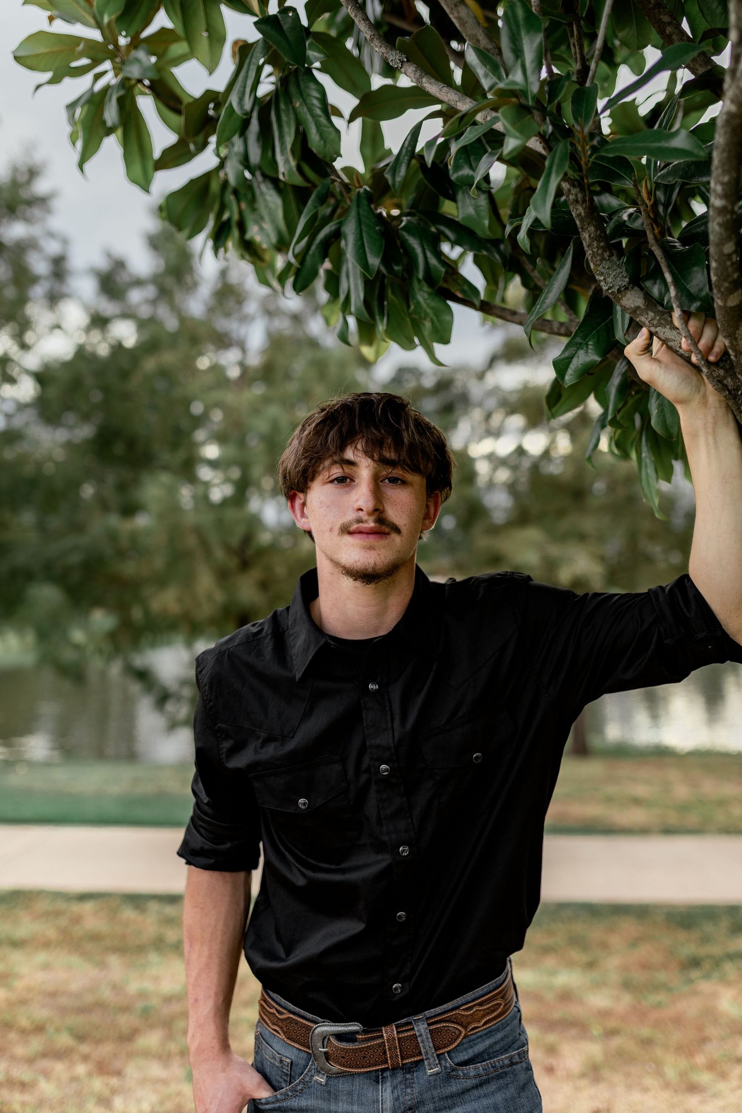 Young man in black shirt leaning against a magnolia tree, posing confidently in an outdoor park setting.