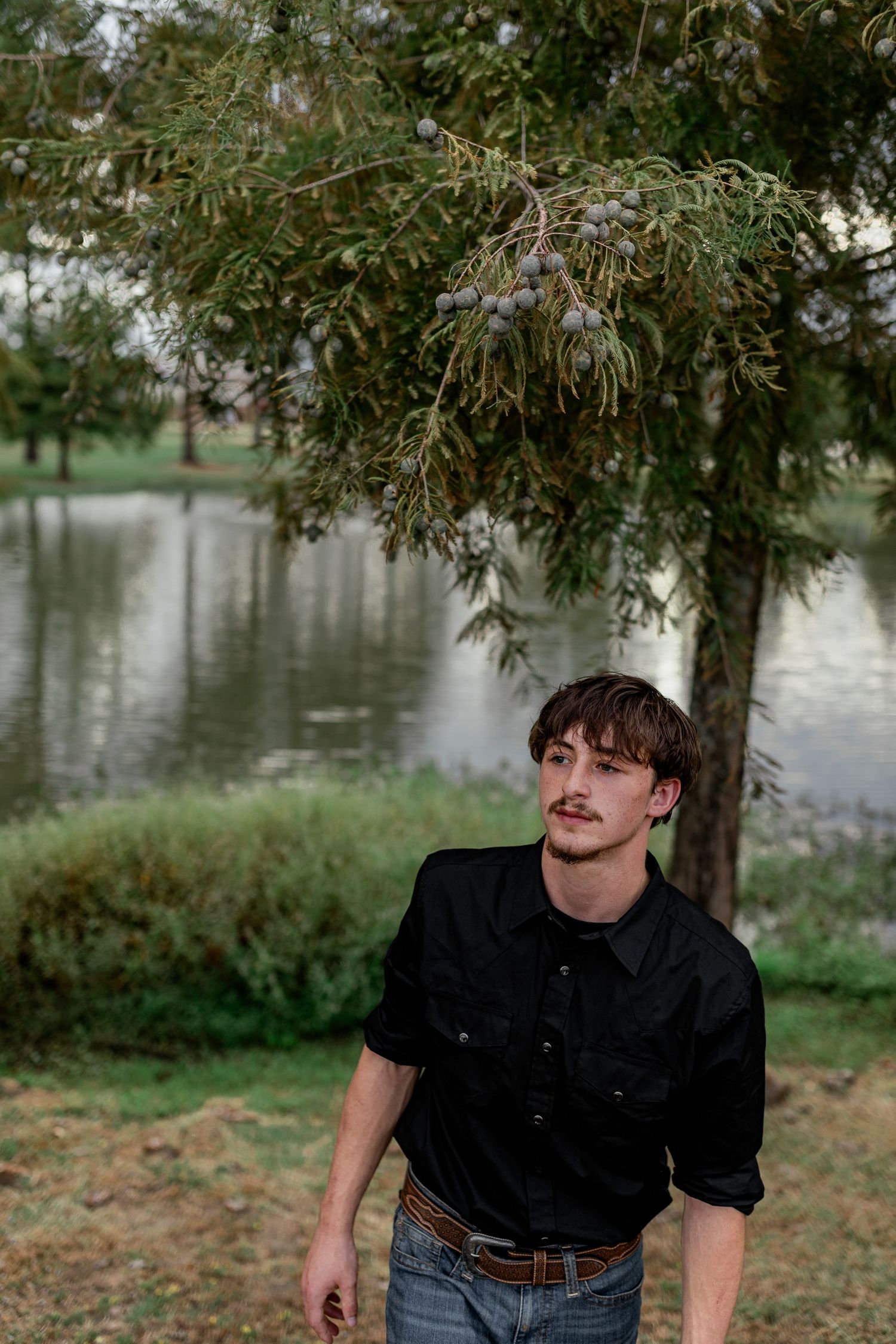 Young man in black shirt standing near a weeping tree beside a calm reflective pond outdoors.
