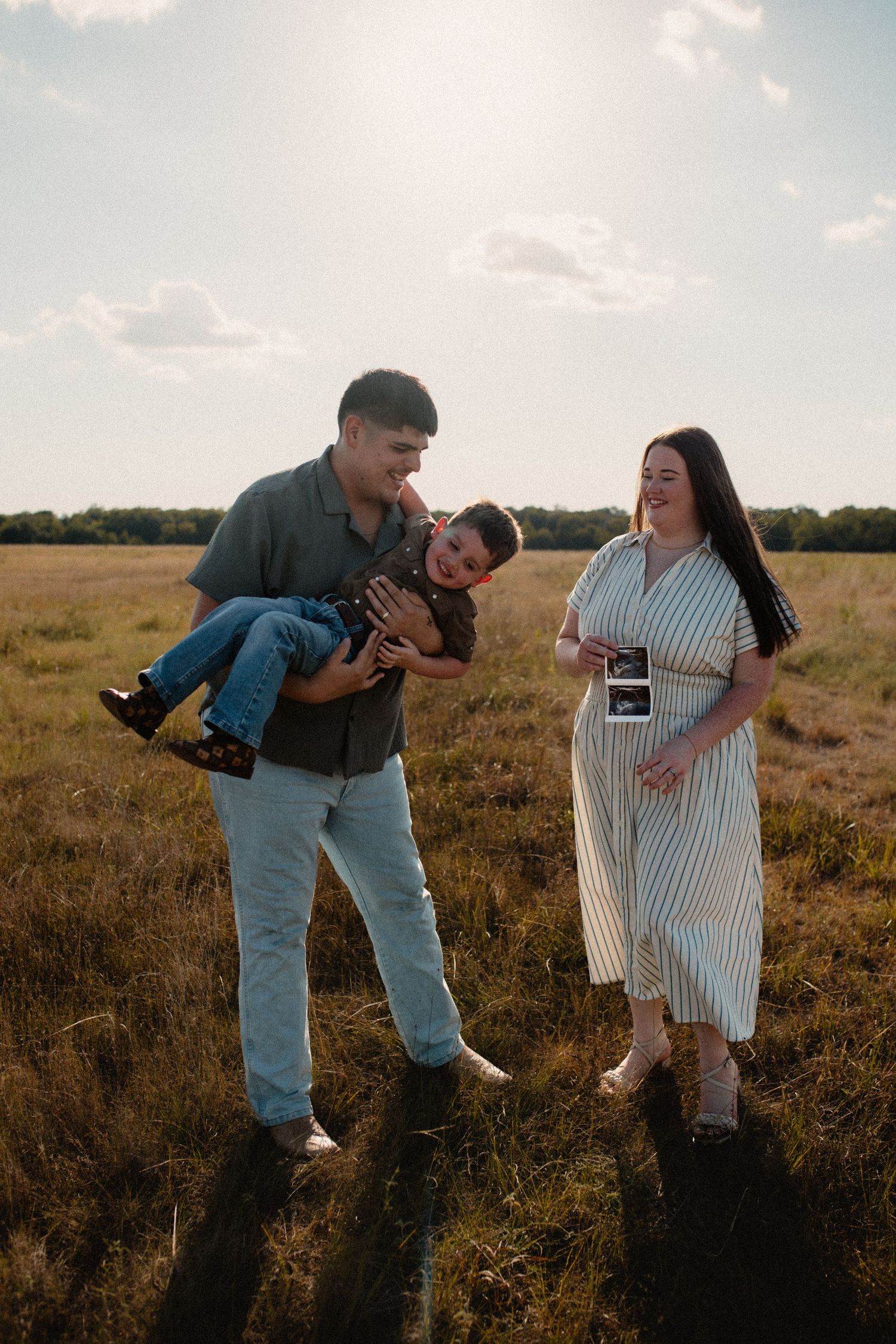 Father holding laughing son while pregnant mother displays ultrasound photo in sunny open field.