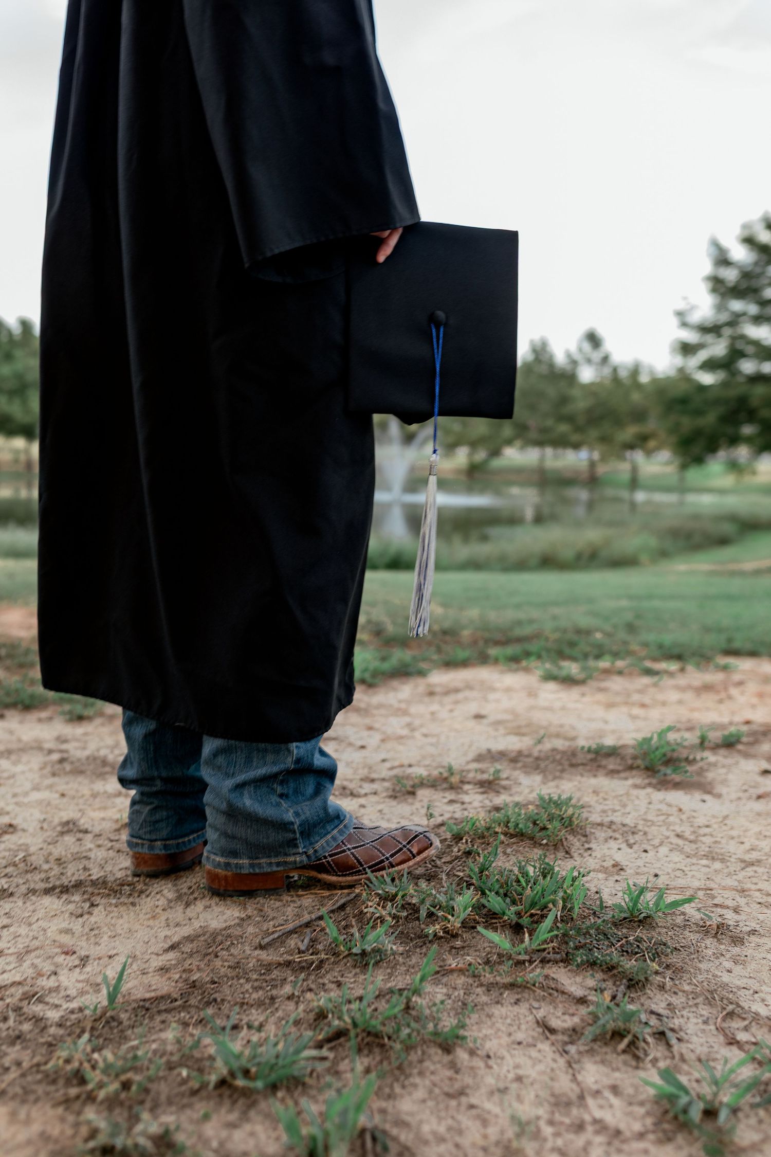 Graduate in black gown holds mortarboard cap while standing on sandy path in casual brown shoes.