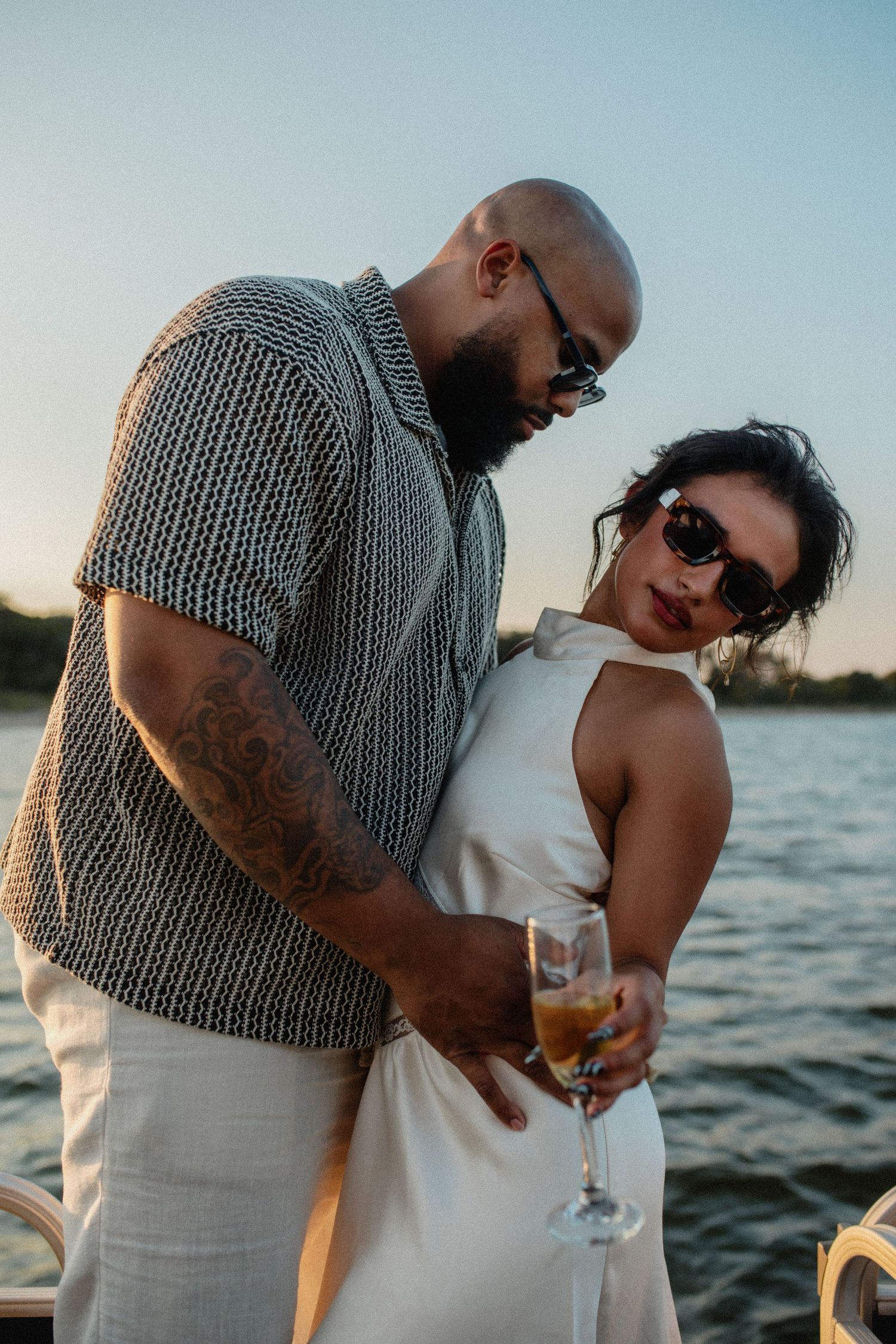 Stylish couple on boat at sunset, bald bearded man holding woman in white dress with champagne.
