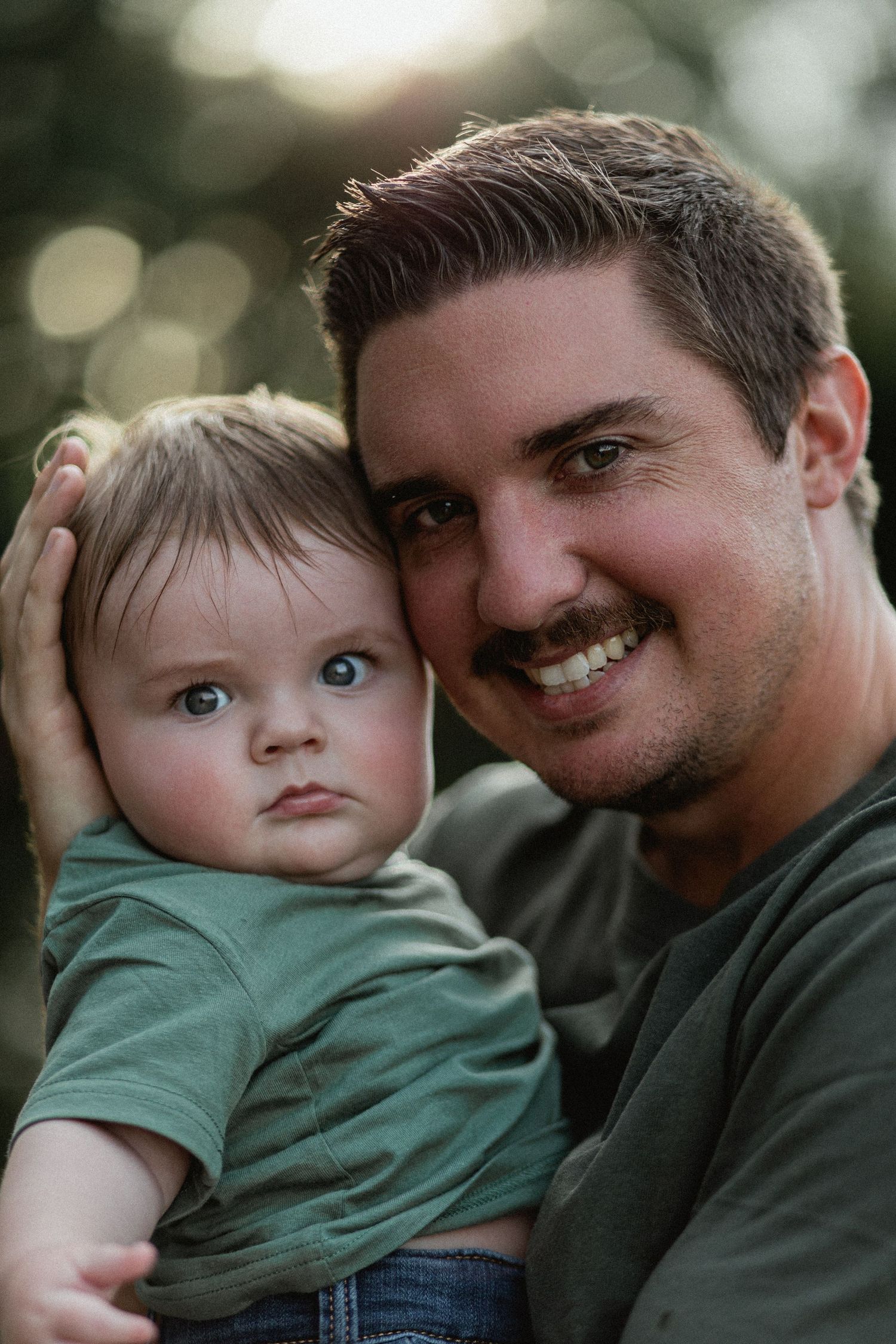 Smiling father holding blue-eyed baby close, cheek to cheek in golden hour light.