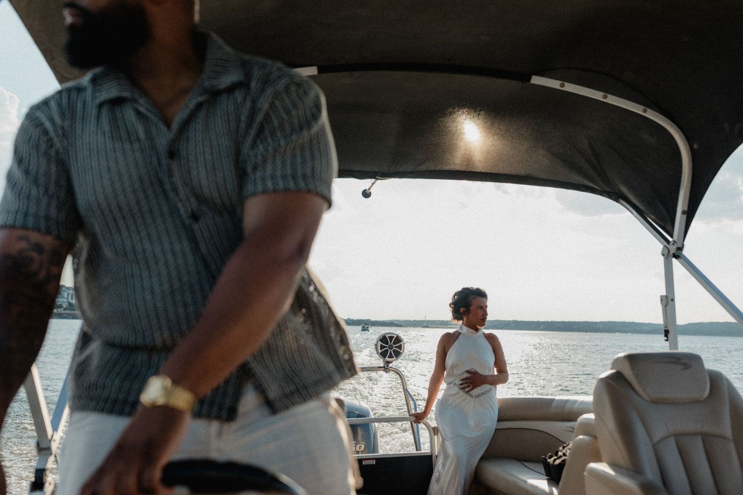 Couple on a pontoon boat on open water, woman in white dress standing near the stern in sunlight.
